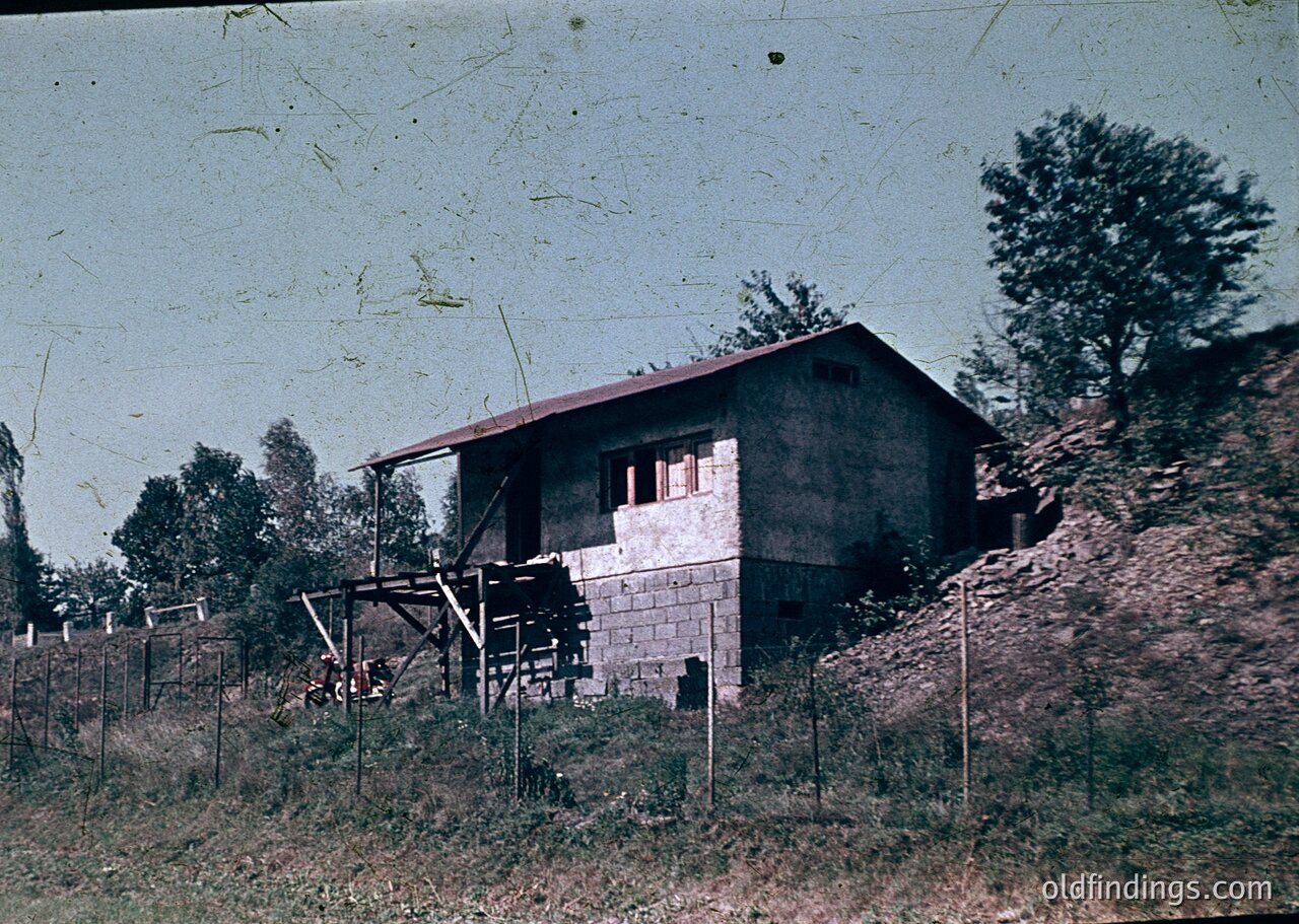 Vintage sepia-toned photo of a single-story concrete house with a pitched roof, set on a sloped hillside. Wooden support beams and a rustic fence frame the property. Lush greenery and trees surround, suggesting a rural or mountainous setting. Likely late 20th century.