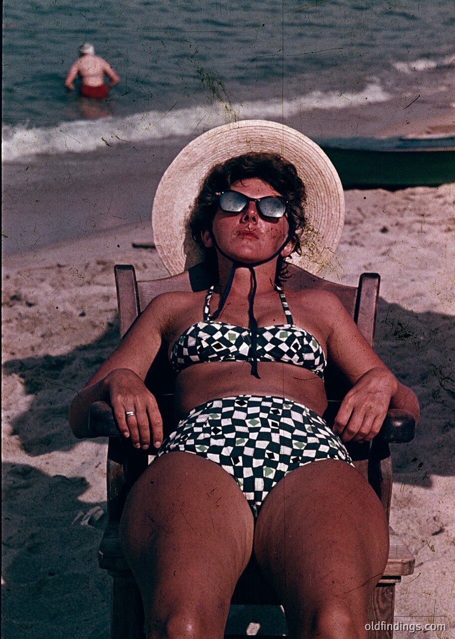 Vintage seaside portrait of a woman in a straw hat and checkered bikini lounging on a wooden deck chair, 1960s beachwear. Sun-bleached sand and calm waters in background with another swimmer. Classic retro coastal lifestyle.