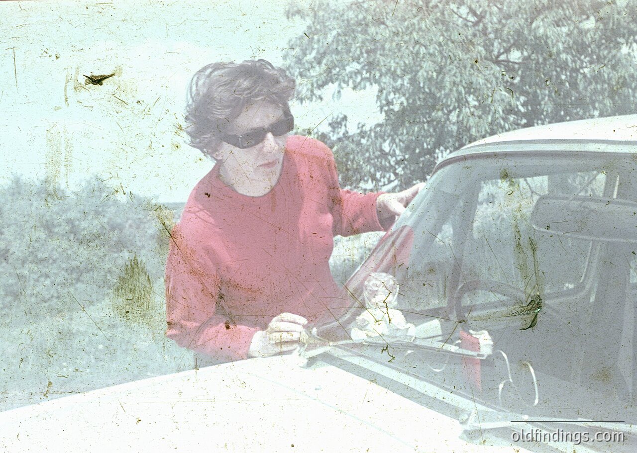 Vintage sepia-toned photo of a woman in a red sweater and sunglasses opening a classic car door, likely a 1960s–1970s model. Rustic outdoor setting with blurred greenery and a dusty road.