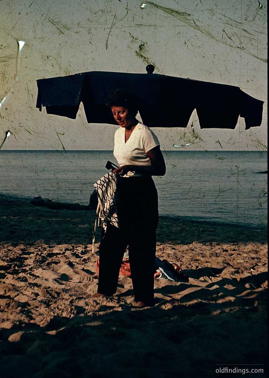 Mid-20th century seaside vendor holding a striped cloth under a large black umbrella, standing on sandy beach near calm waters. Clothing suggests 1950s–1960s fashion (, , , , ).
