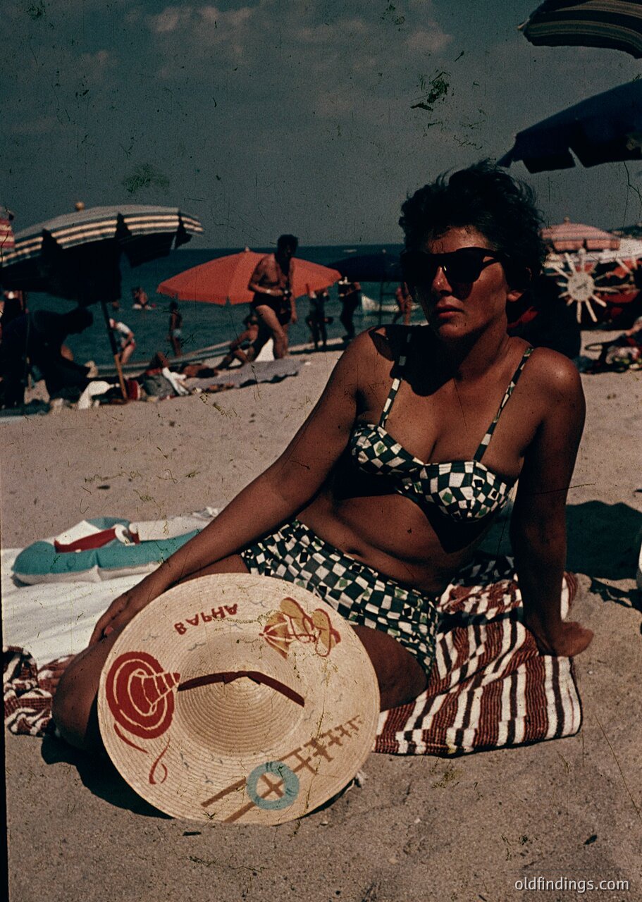 Vintage seaside portrait from the 1960s-70s, featuring a woman in a striped swimsuit and straw hat with floral design. She sits on sandy beach near umbrellas and towels, with ocean and beachgoers in background. Classic retro fashion and mid-century beach culture.