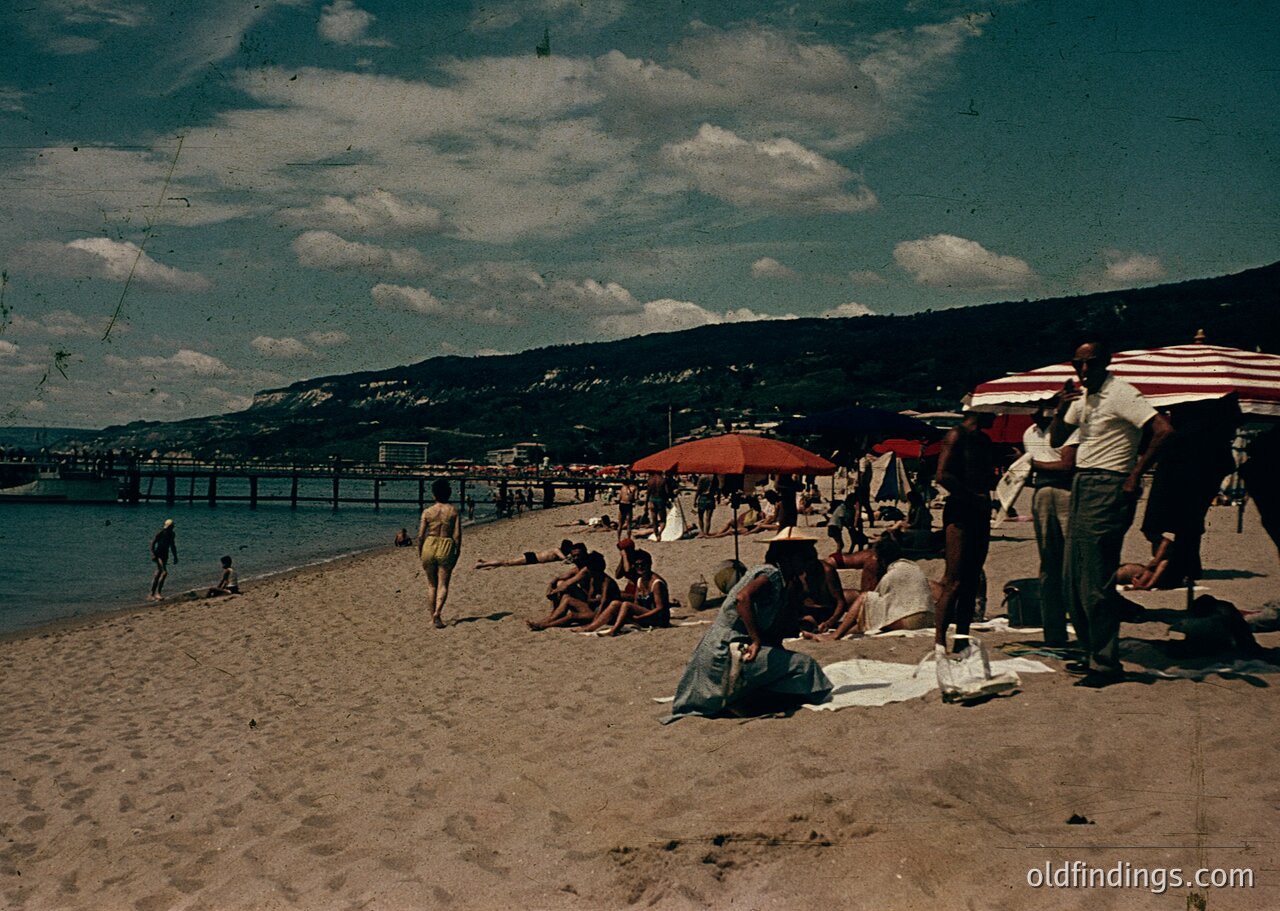 Vintage seaside scene with wooden pier extending into calm waters. Crowded sandy beach with mid-20th-century attire—swimsuits, suits, and wide-brim hats. Red/white striped umbrellas provide shade. Hilly landscape in background. Likely European coastal resort, 1950s-60s.
