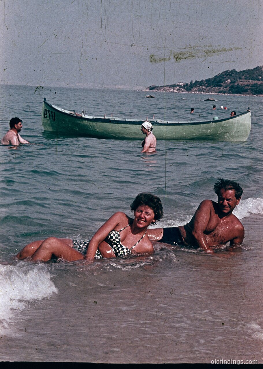 Mid-20th century seaside scene: Couple in wet swimwear lounges on shallow waves, framed by a green wooden boat labeled "PNH" and rocky coastline. Casual beachgoers swim nearby.