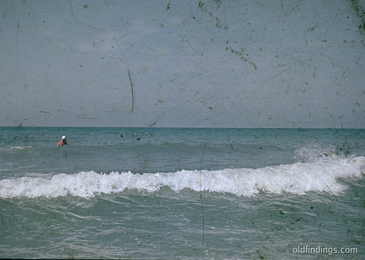 Vintage seaside shot featuring a lone swimmer in mid-wave. Faded, textured print suggests mid-20th century coastal photography. Waves crash against a rocky shoreline; distant beach appears deserted. Ideal for historical research or nostalgic design references.