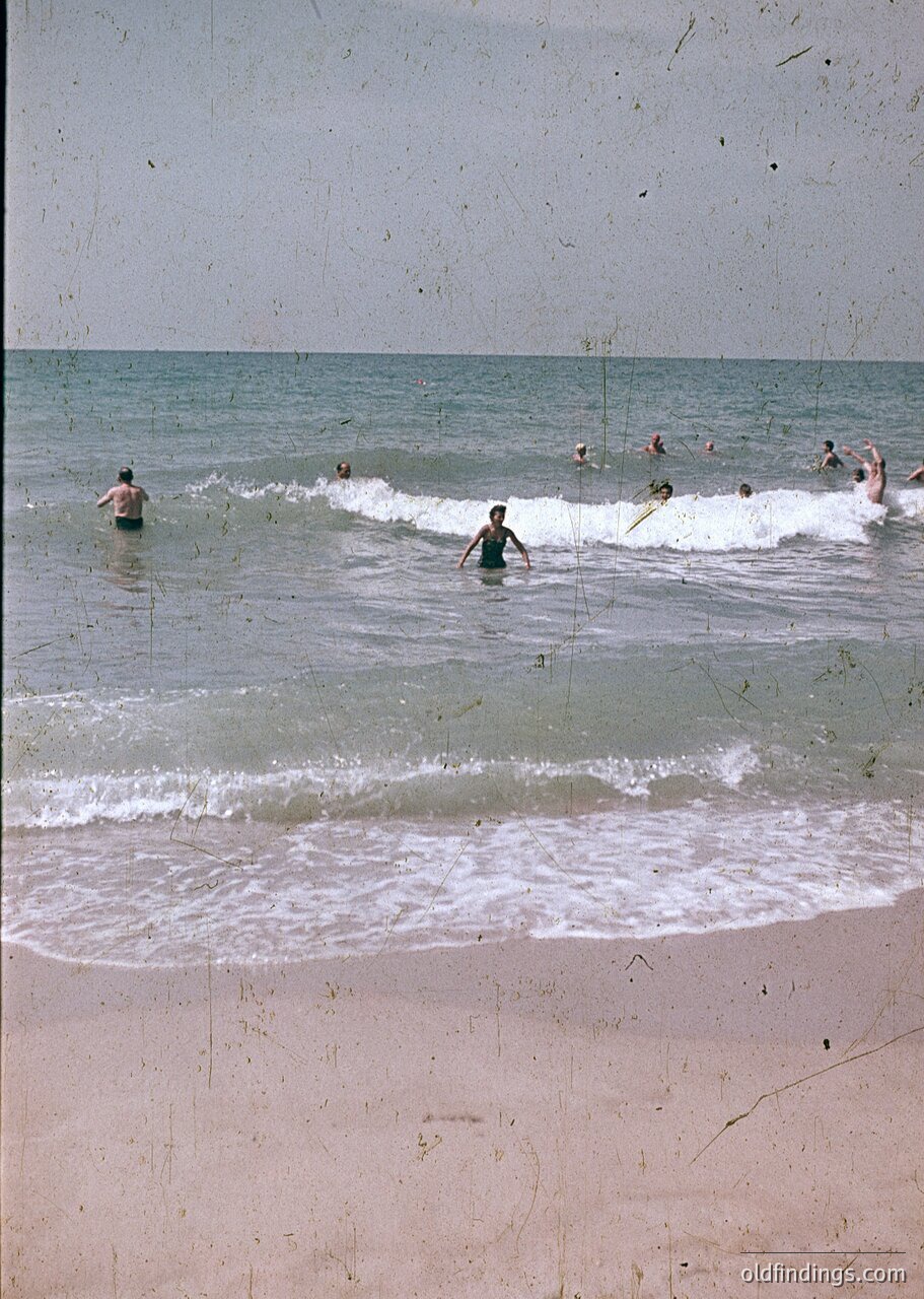 Vintage seaside scene with 1960s-style swimwear, groups wading in shallow waves. Beach appears sandy with visible footprints. Overcast sky suggests mid-century coastal tourism. Potential or seaside vibe.