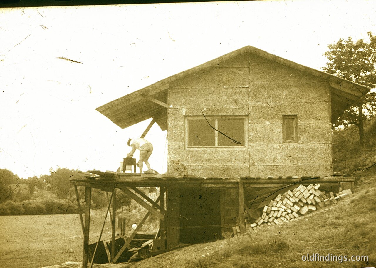 Vintage sepia-toned photo of a small, elevated wooden guardhouse with a peaked roof, likely a 20th-century border or railway lookout. A lone figure in period attire stands on a platform, holding a rifle. Stacked bricks and wooden supports suggest construction or maintenance activity. Rustic, rural setting with blurred greenery in background.