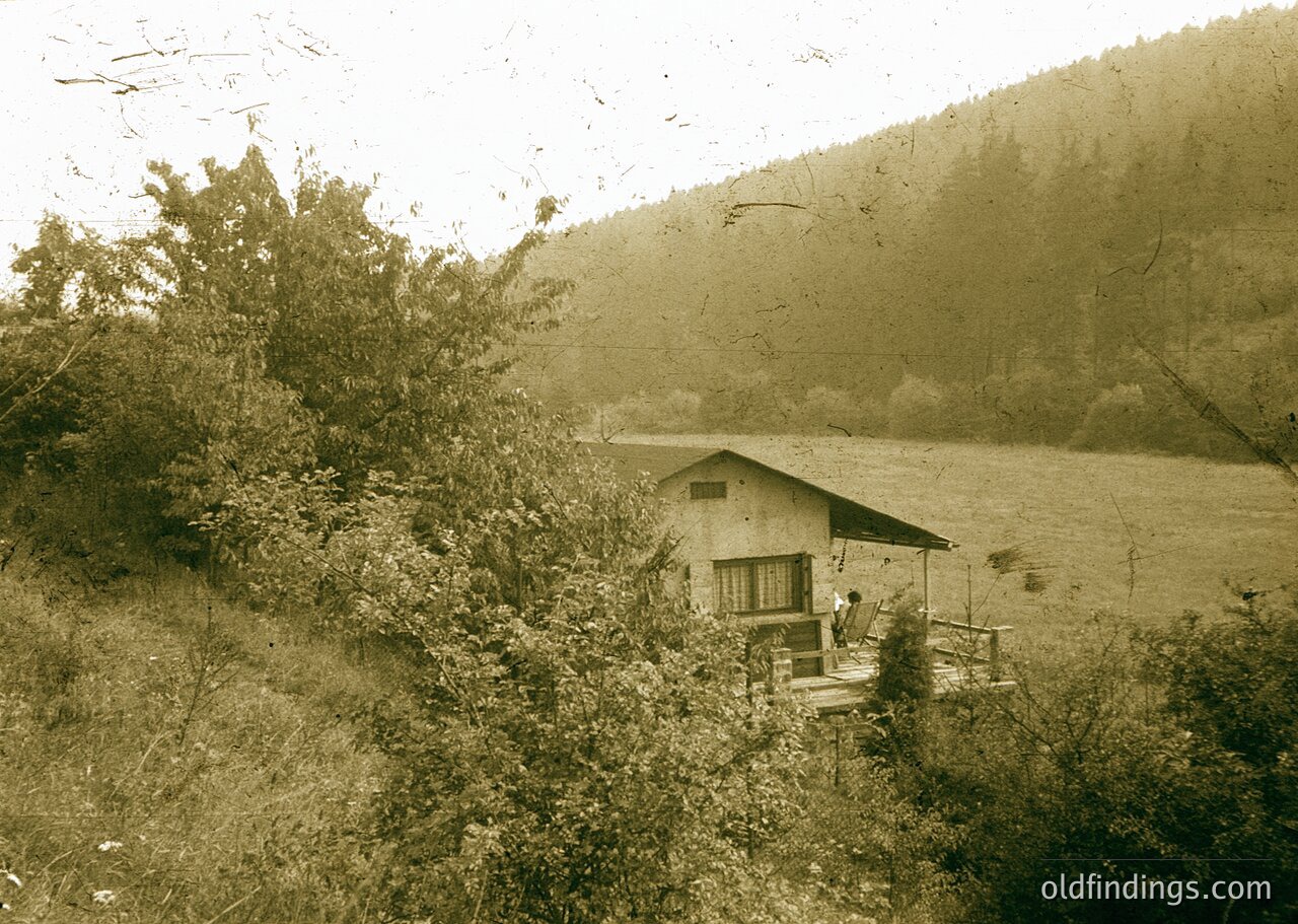 Vintage sepia-toned rural scene featuring a small wooden cabin with a porch overlooking a lake, surrounded by dense shrubbery and hills. Likely early-to-mid 20th century.