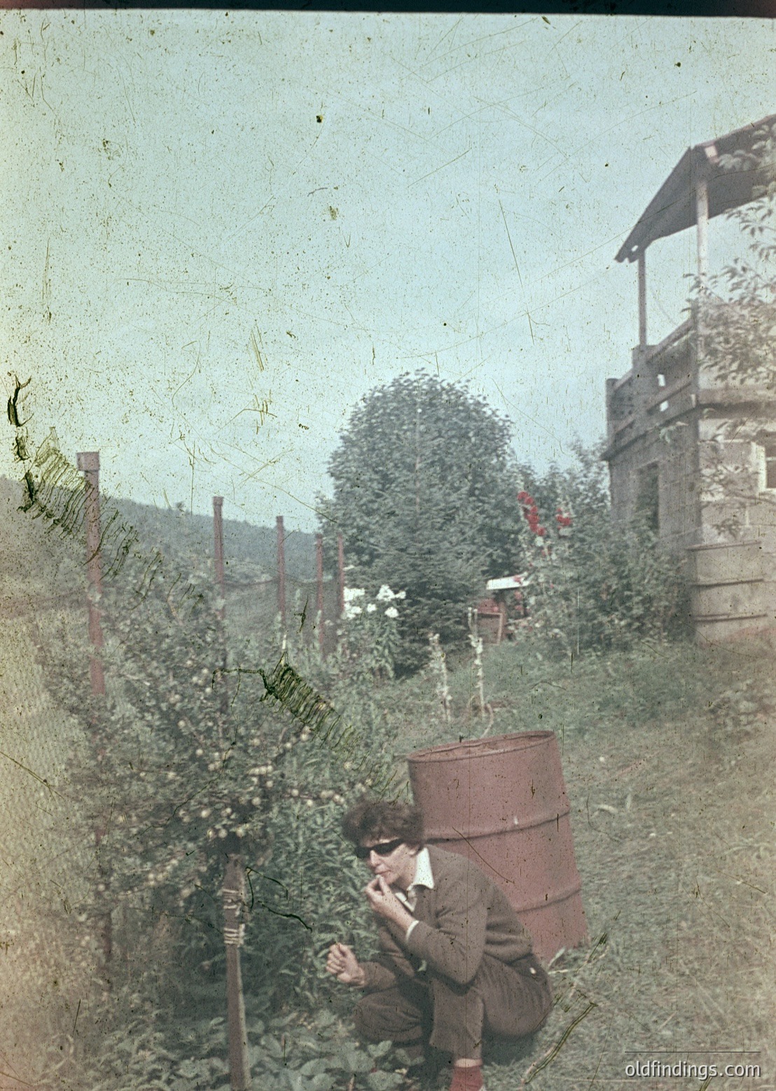 Vintage sepia-toned photo of a person kneeling in a lush garden, using an early 20th-century telephone. Wooden fence, rustic barrel, and overgrown greenery frame the scene. Likely rural, early .