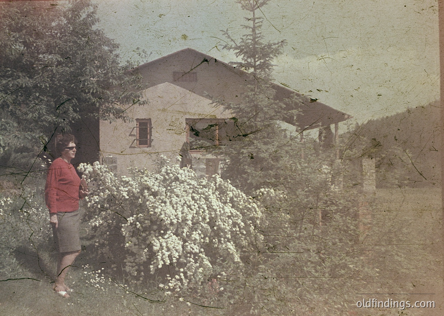 Vintage sepia-toned photo of a woman in a red blouse and shorts posing beside a blooming tree, likely a peach or plum, in front of a modest, two-story house with a pitched roof. The structure appears weathered, with visible cracks and aged plaster. The setting suggests a rural or suburban area, possibly early-to-mid 20th century.