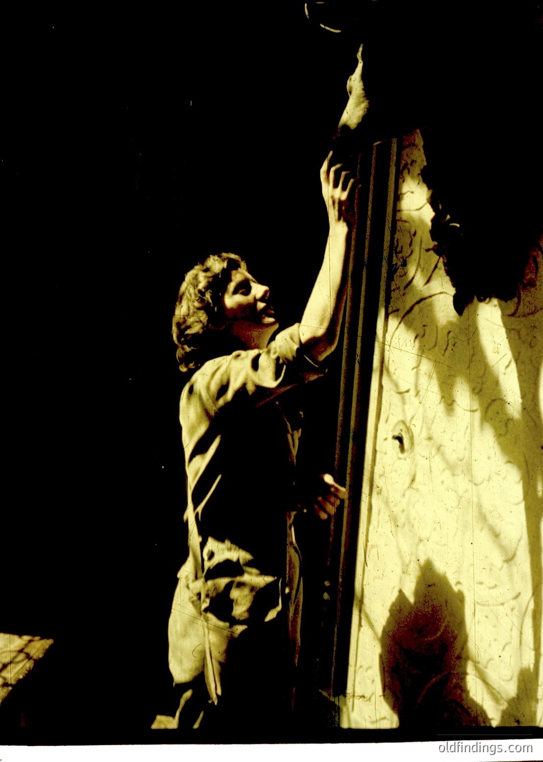Black-and-white statue of a young boy reaching upward, gripping a vertical pole. The figure appears to be in motion, possibly climbing or ascending. The background shows a draped, semi-transparent cloth with faint floral or geometric patterns. Likely a 20th-century theatrical or cinematic prop, evoking 1950s–1960s drama.