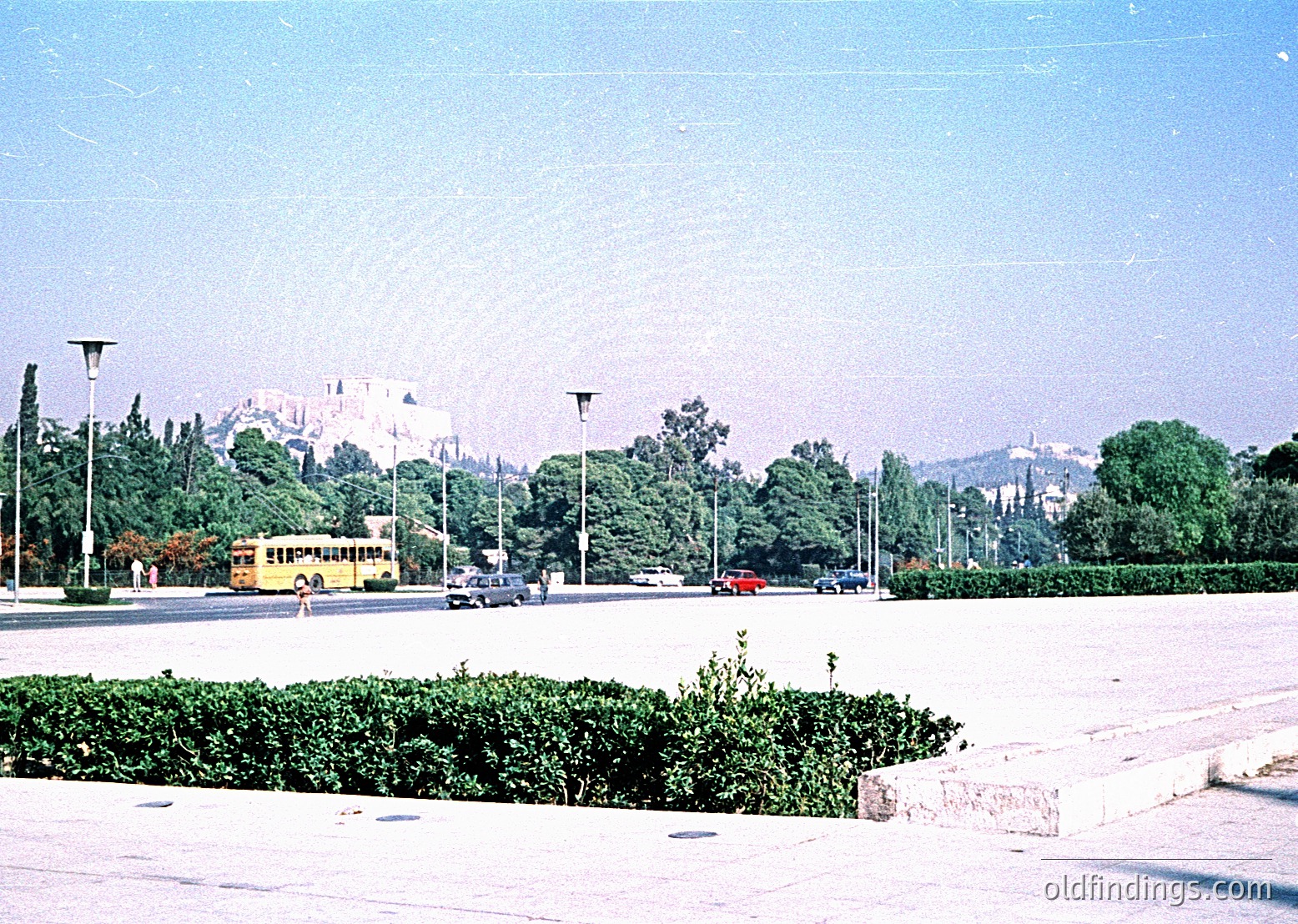 Vintage urban scene featuring a yellow bus and classic cars on a wide, tree-lined boulevard. Prominent historic castle ruins in background, likely . Well-maintained hedges and vintage street lamps enhance mid-century urban design.