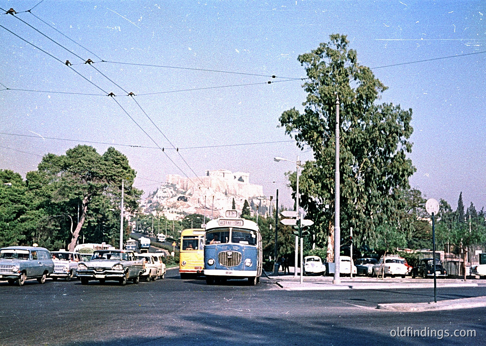 Vintage 1950s-60s urban scene featuring a blue & yellow trolleybus at a busy intersection. Classic cars, overhead wires, and lush greenery frame a mid-century cityscape. Likely North American due to vehicle design.