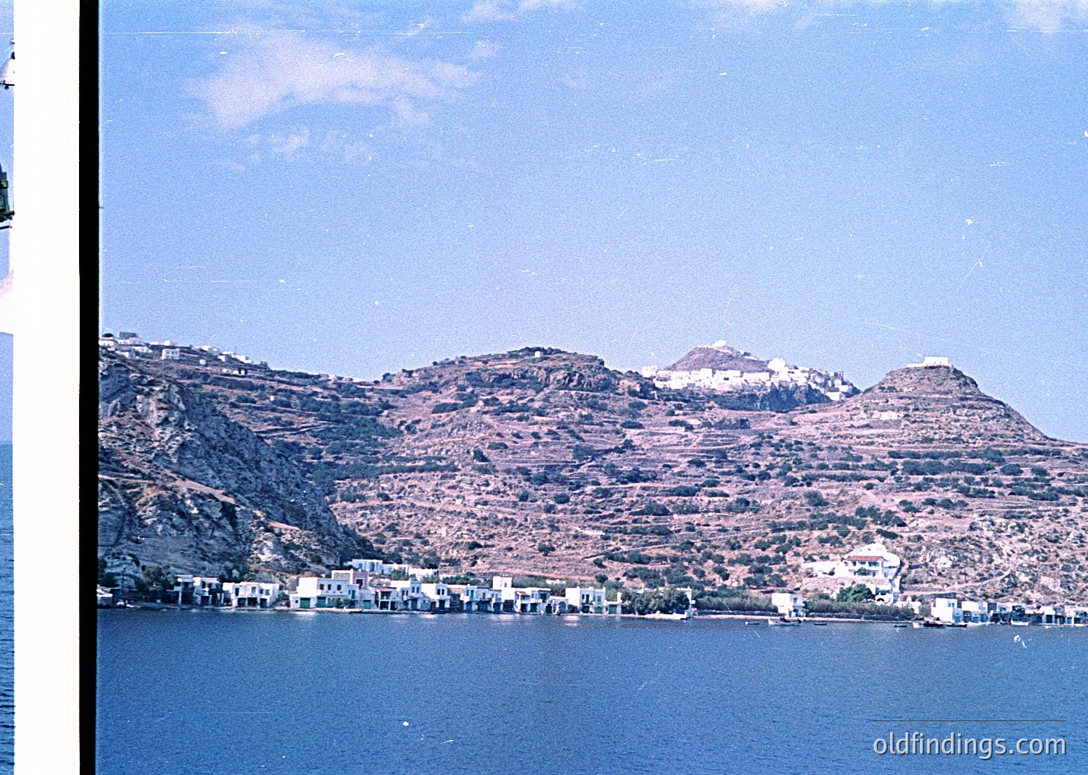 Coastal village nestled between rugged hills and deep blue waters, likely Mediterranean. Whitewashed buildings with flat roofs line the shore, framed by terraced slopes. Mid-20th century (1950s–1960s) color photo, suggesting post-war reconstruction or early tourism development.