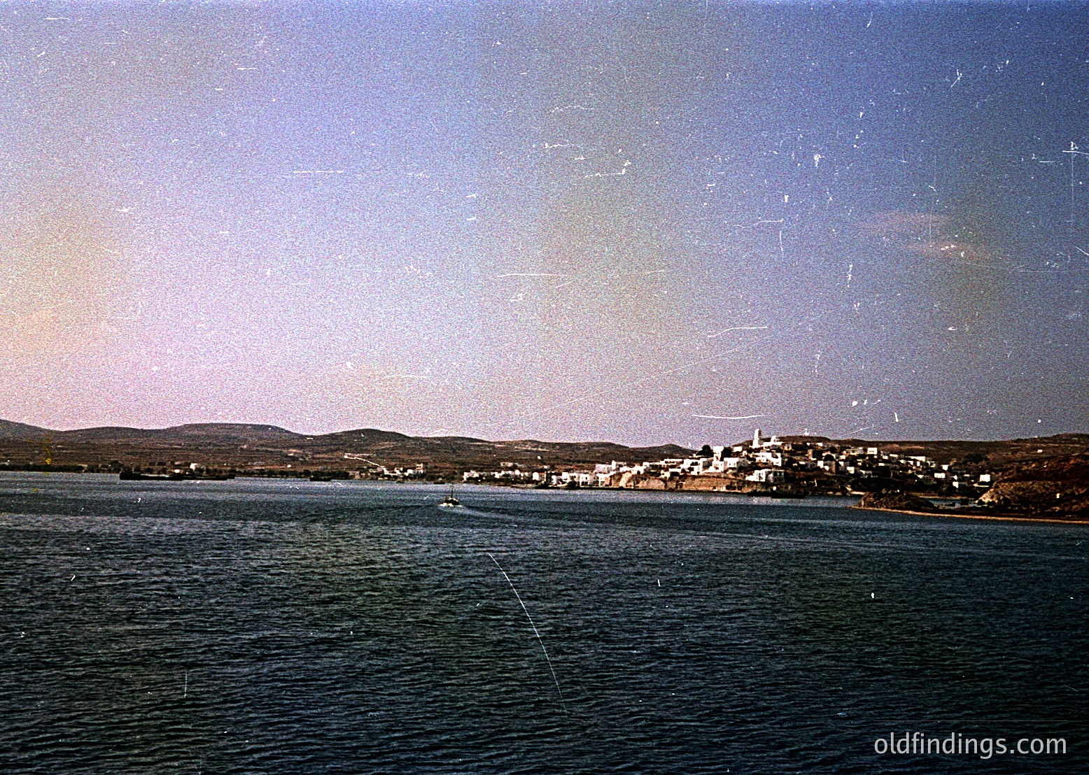 Vintage coastal town framed by calm waters, likely Mediterranean. Low-lying buildings with flat roofs and whitewashed walls line the shoreline under a hazy sky. Distinctive lighthouse on a hill to the right. Sepia-toned, suggesting 19th–early 20th century. Ideal for historical research or vintage travel inspiration.