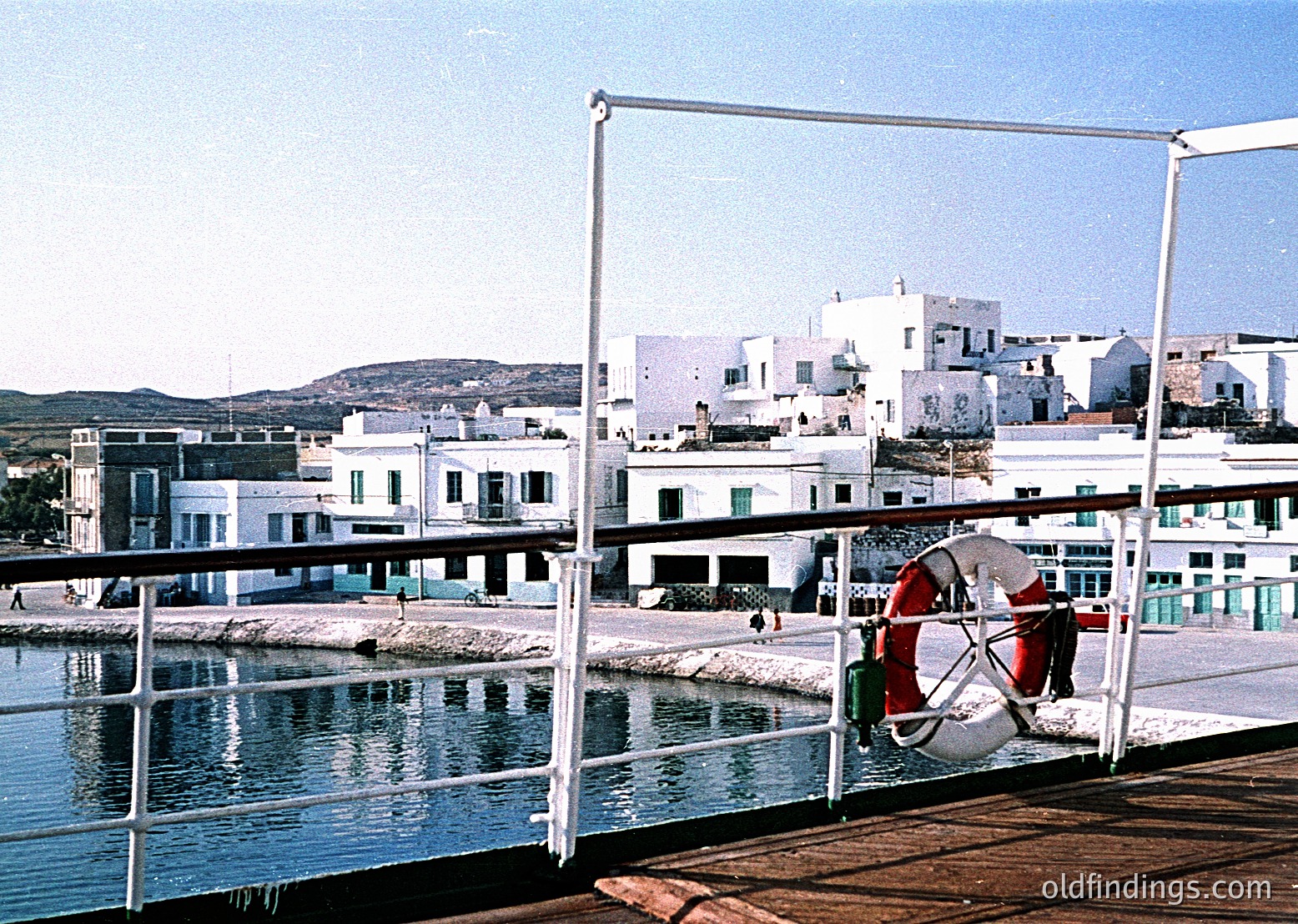 Mid-20th century seaside town with whitewashed Mediterranean architecture. Prominent lifebuoy on metal railing by calm harbor water. Low-rise buildings with flat roofs and balconies, framed by distant hills under clear skies. Likely or style.