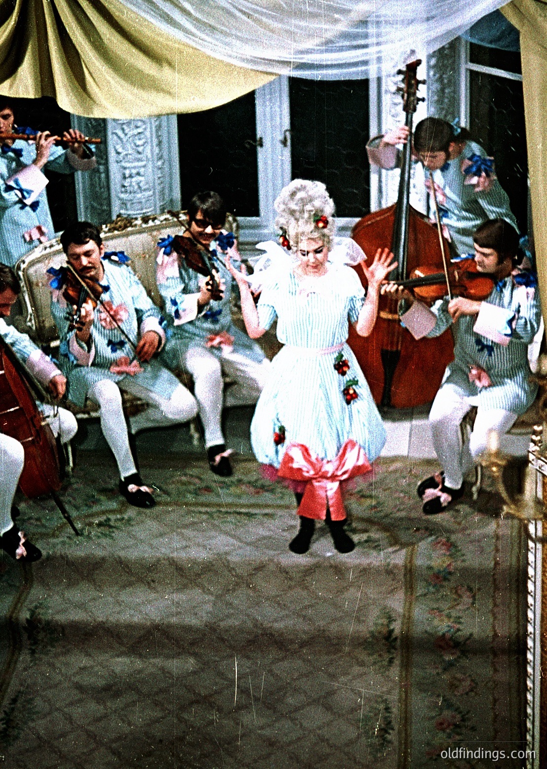 Vintage 19th-century indoor musical performance with classical string ensemble. Central figure in elaborate white gown with red accents dances, surrounded by musicians in period attire—violinists, cellists, and a harpist. Ornate interior with draped curtains and decorative moldings. Evokes European ballroom culture.
