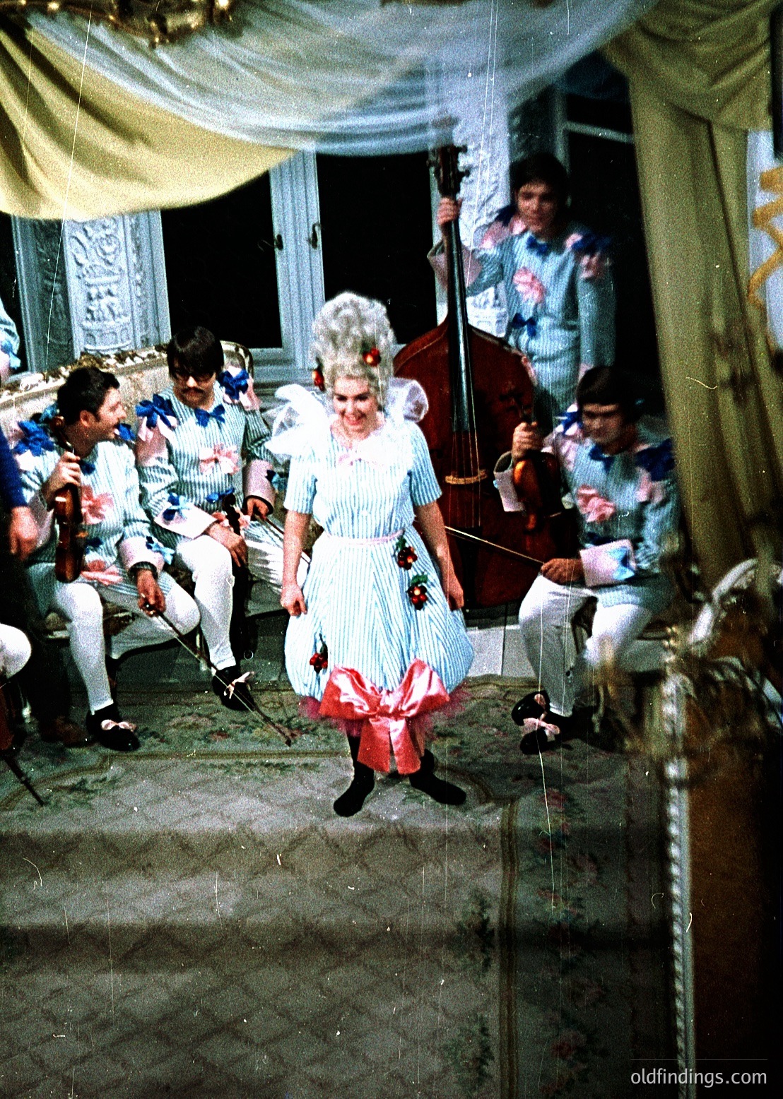 Vintage stage performance featuring a woman in a 19th-century ballgown with floral embroidery, white gloves, and a decorative wig, dancing on a wooden platform. Surrounding musicians in striped shirts and white pants play string instruments. Ornate ceiling and draped curtains suggest a theatrical or opera house setting, likely late 19th to early 20th century.