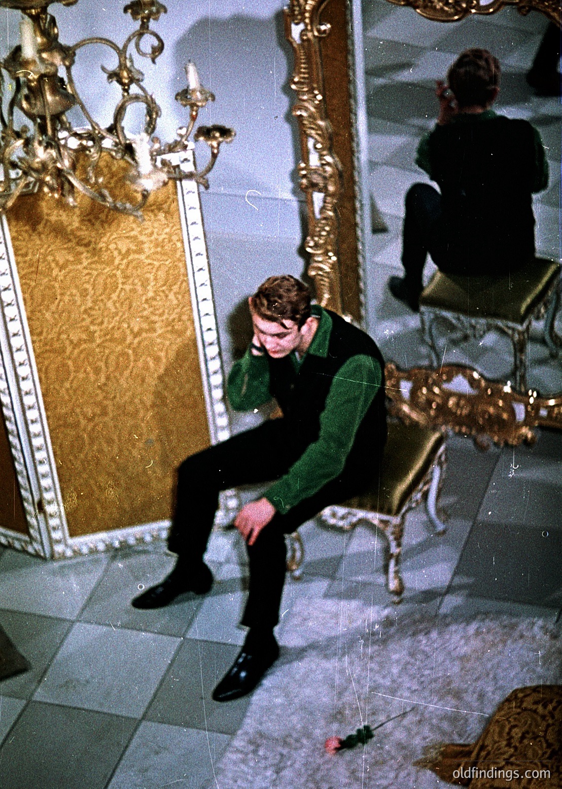 Young man in 1960s-70s streetwear (green sweater, dark pants, boots) sits on ornate marble floor in a grand, mirrored room. Gold-framed mirrors and vintage chandelier reflect opulent interiors. Possible Eastern Bloc-era architecture or luxury hotel lobby.