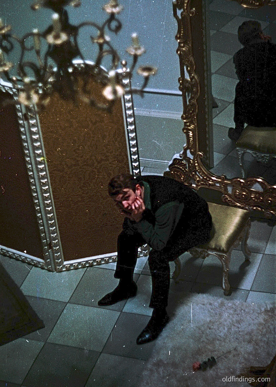 A man in a dark, tailored suit sits on an ornate, gold-framed chair in a grand, mirrored room. Reflections reveal a chandelier and classical interior. Likely 1950s–1970s European interior design.