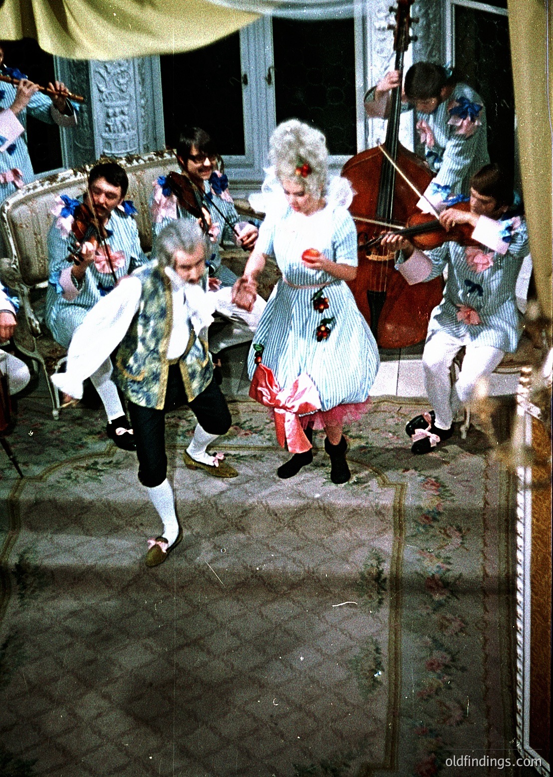 Vintage indoor dance scene with 19th-century-inspired costumes: men in striped pants with vests, women in puff-sleeve dresses with floral bows. Violinist and cellist play in background. Ornate wooden furniture and chandelier suggest a grand ballroom. Likely late 19th to early 20th century European social event.