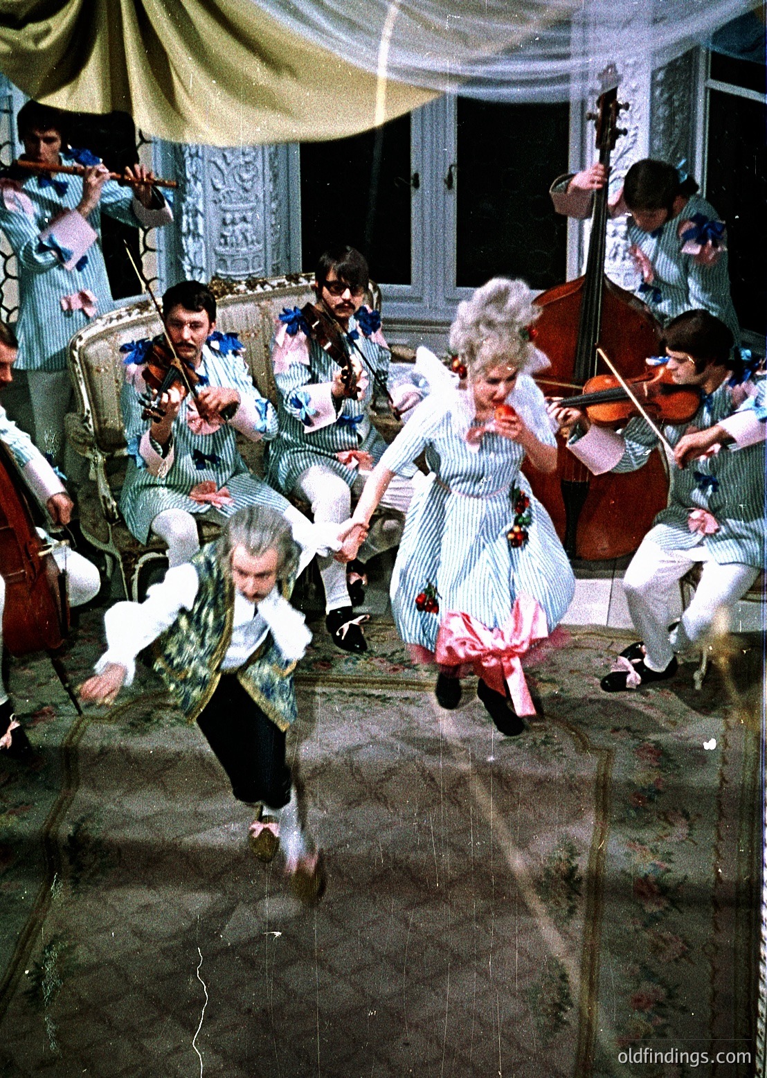 Vintage 1970s indoor folk dance with musicians playing traditional instruments—violins, accordion, cello. Couple in mid-dance, woman in floral dress with white apron, man in patterned vest and white shirt. Orchestra seated on ornate chairs, framed by draped curtains and classical architecture. Authentic cultural celebration.