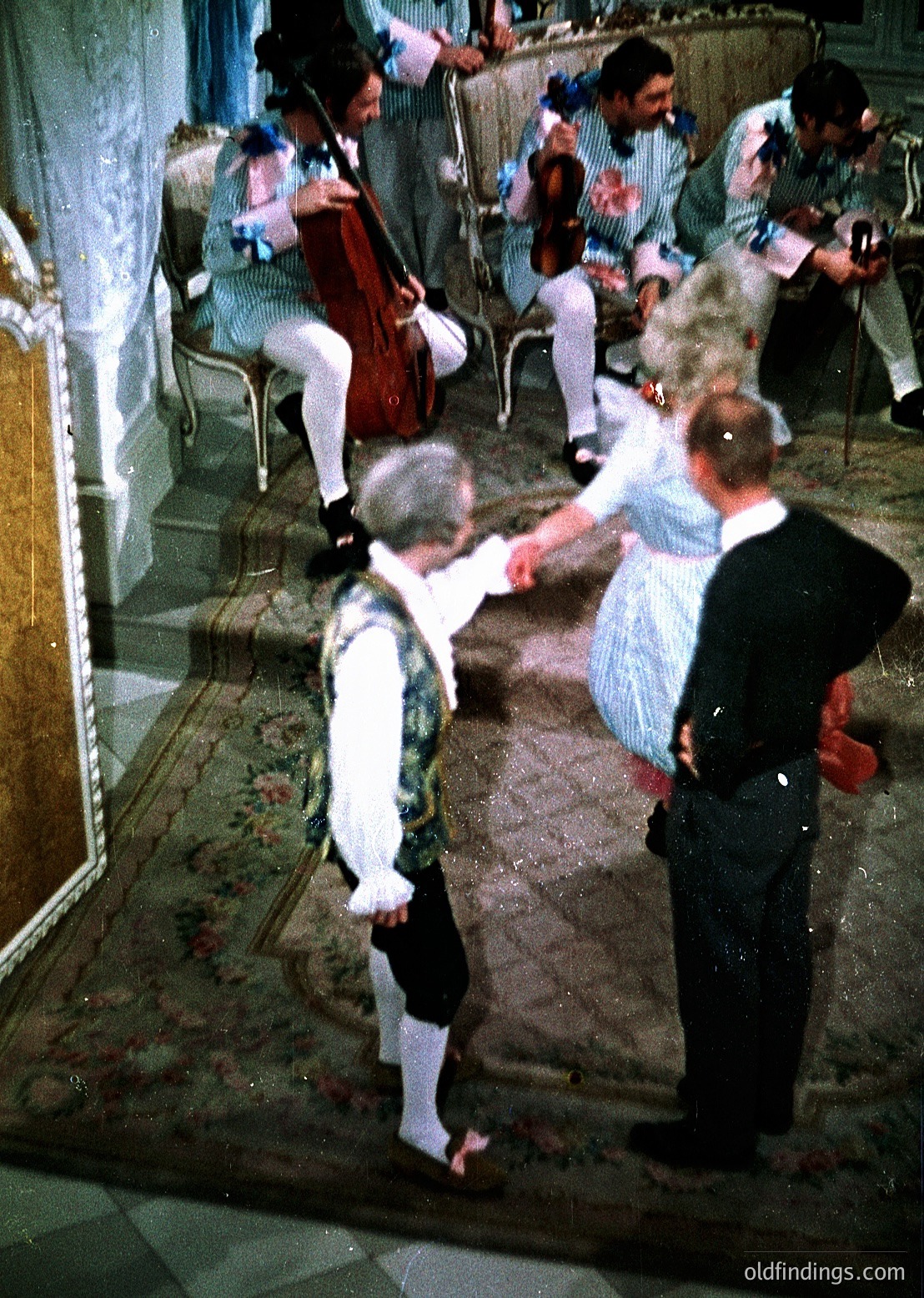Vintage ballroom scene featuring classical attire: a man in 18th-century formal wear (white breeches, vest, stockings) dancing with a woman in a light blue dress. Orchestra plays in background with musicians in period costumes. Ornate interior with wooden paneling and chandelier. Likely European, 18th-century ballroom setting.