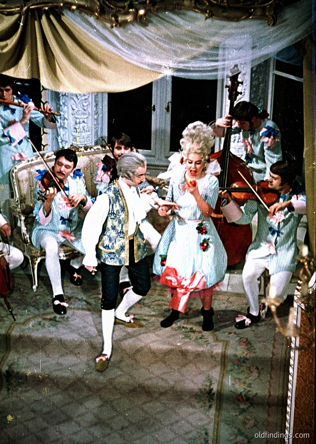 19th-century costume ballroom scene with string quartet performing. Men in tailored white coats with cravats, women in elaborate gowns with floral accents. Ornate chandeliers and draped curtains frame the setting. Likely European aristocratic event, 1850s-1870s.