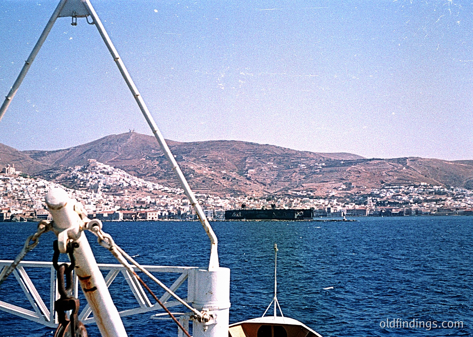 Vintage seascape shot from a boat’s rail, showing a coastal town nestled between hills and deep blue waters. Mid-20th century architecture with whitewashed buildings and a prominent church spire.