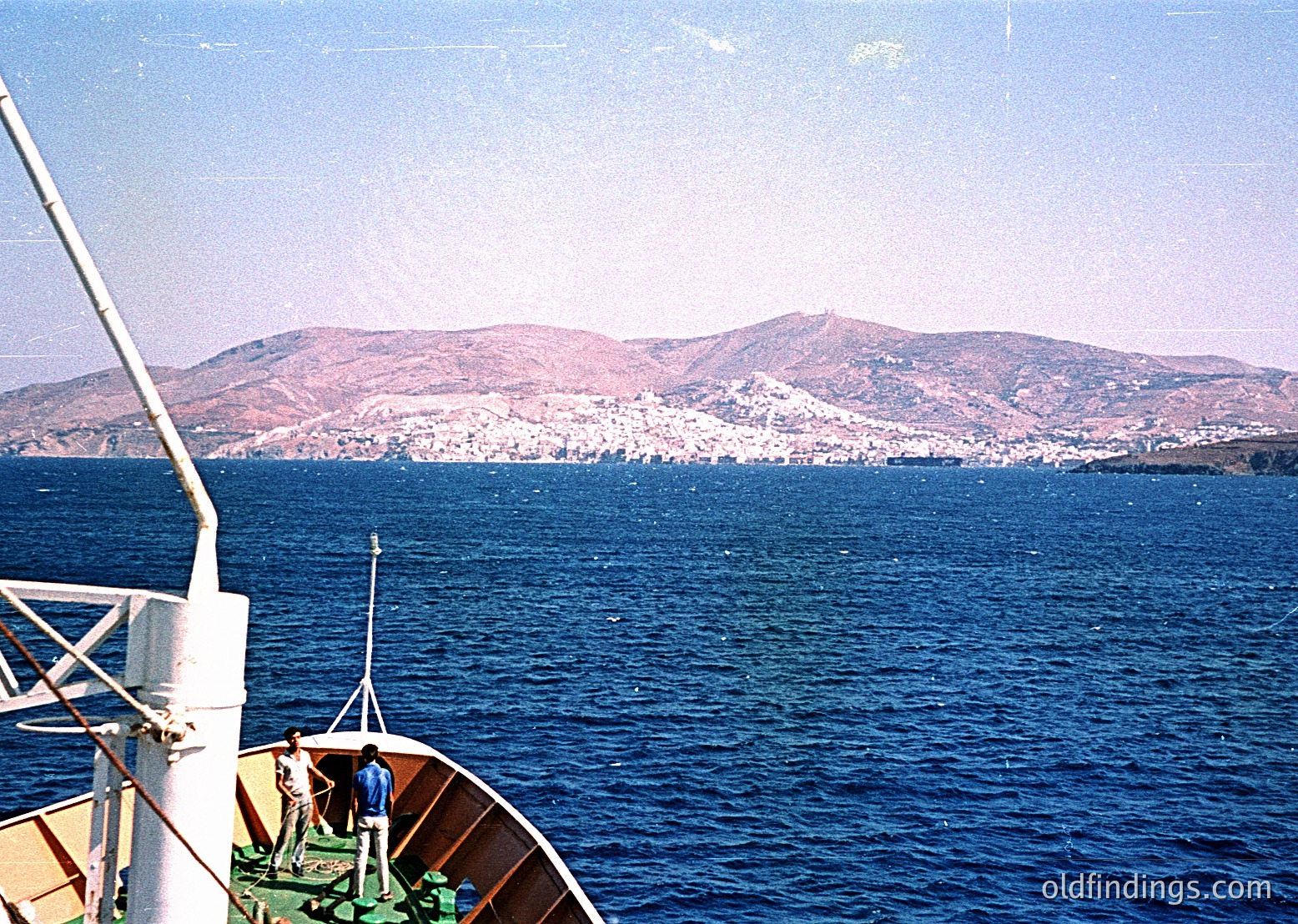 Vintage boat view of coastal cityscape with rolling hills in background. Mid-20th century seaside scene, likely Mediterranean. Distinctive low-angle perspective from vessel deck.