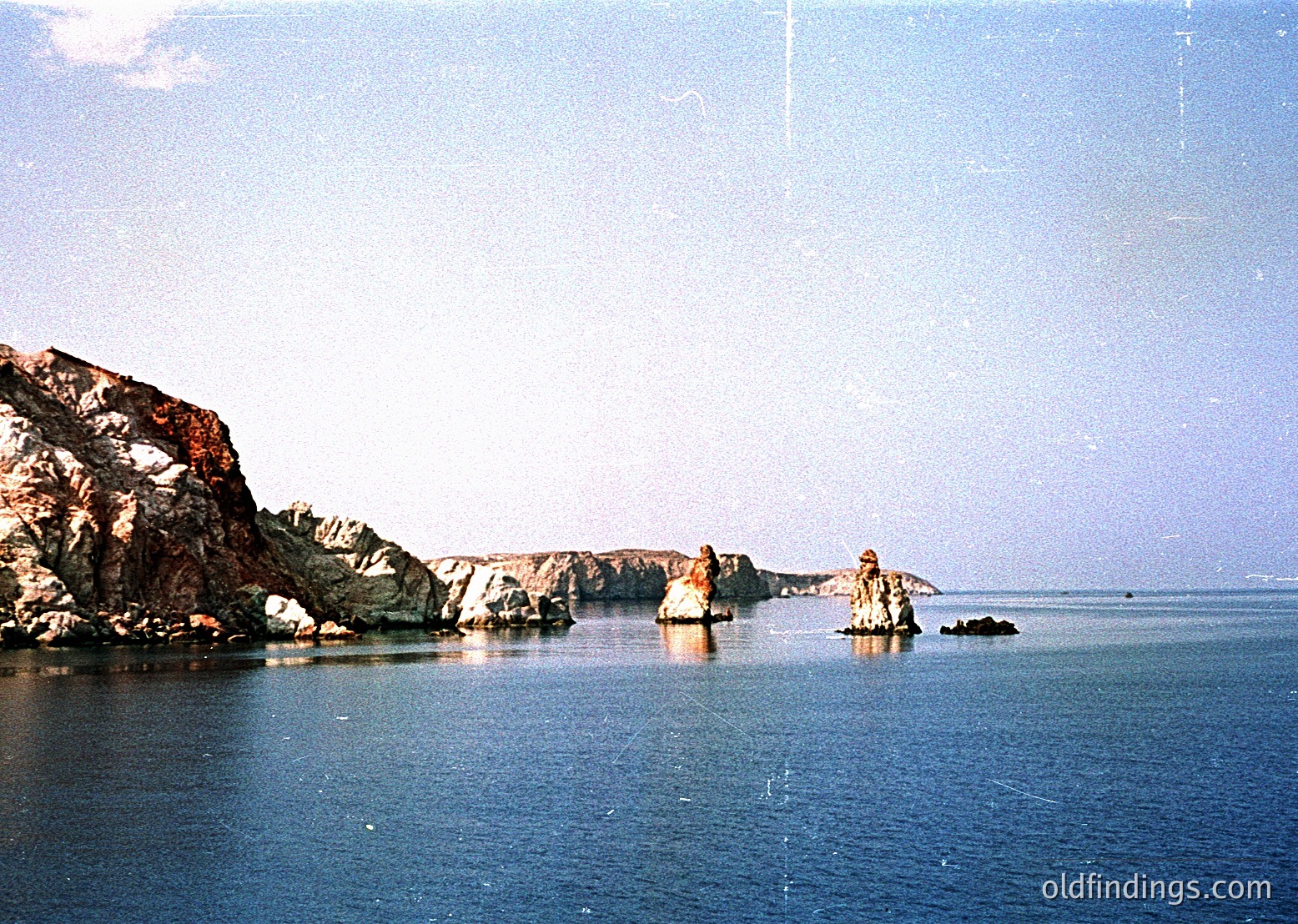 Vintage coastal scene featuring jagged volcanic rock formations rising from calm, reflective waters. Low-angle sunlight casts subtle highlights on the rocks. Likely color film due to sepia-toned aesthetic. Ideal for , , and references.