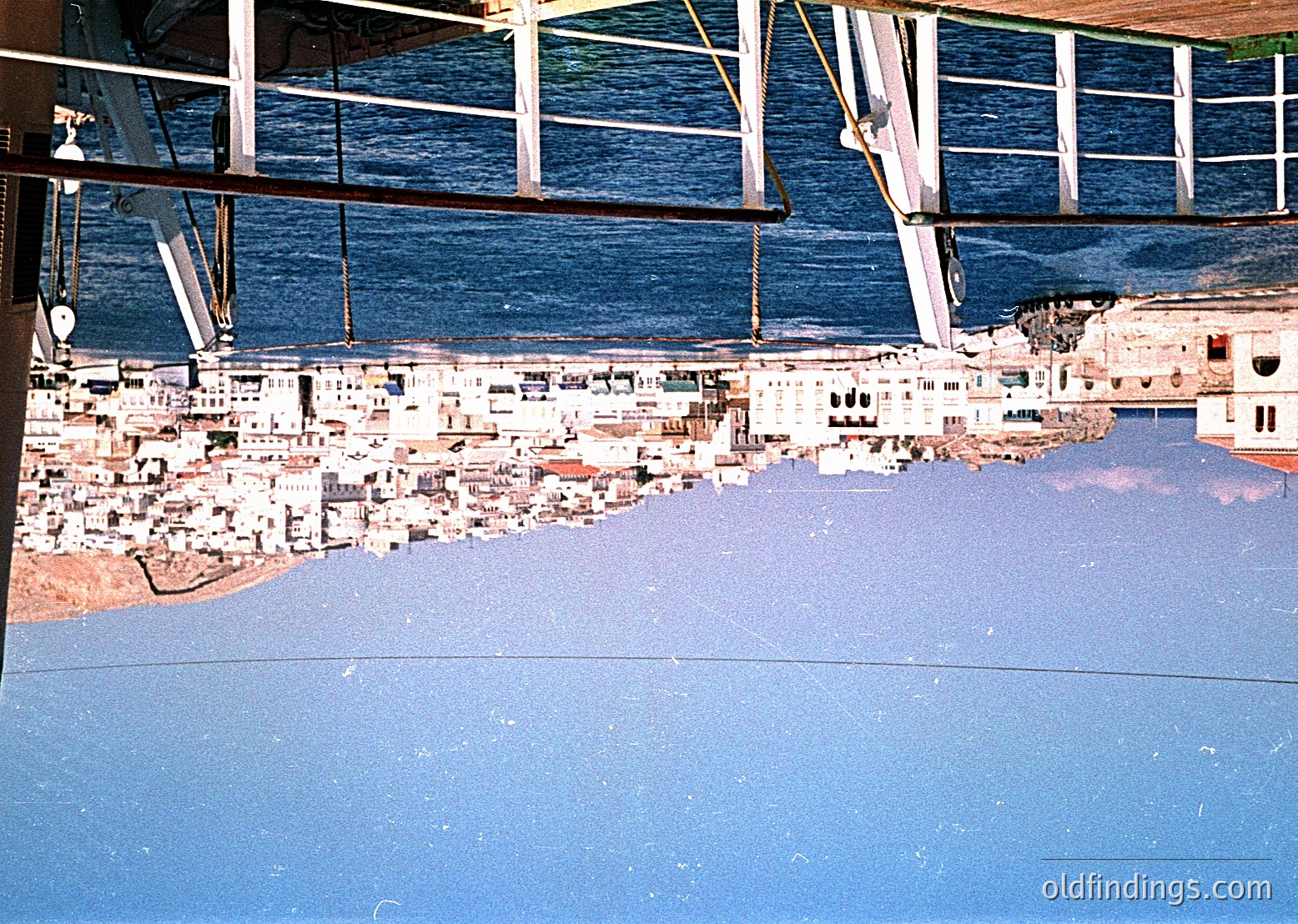 Aerial view of coastal cityscape reflected in ship’s hull, likely Mediterranean. Dense urban layout with narrow streets and light-colored buildings. Prominent pier extends into deep blue waters, with ship railings framing the scene.
