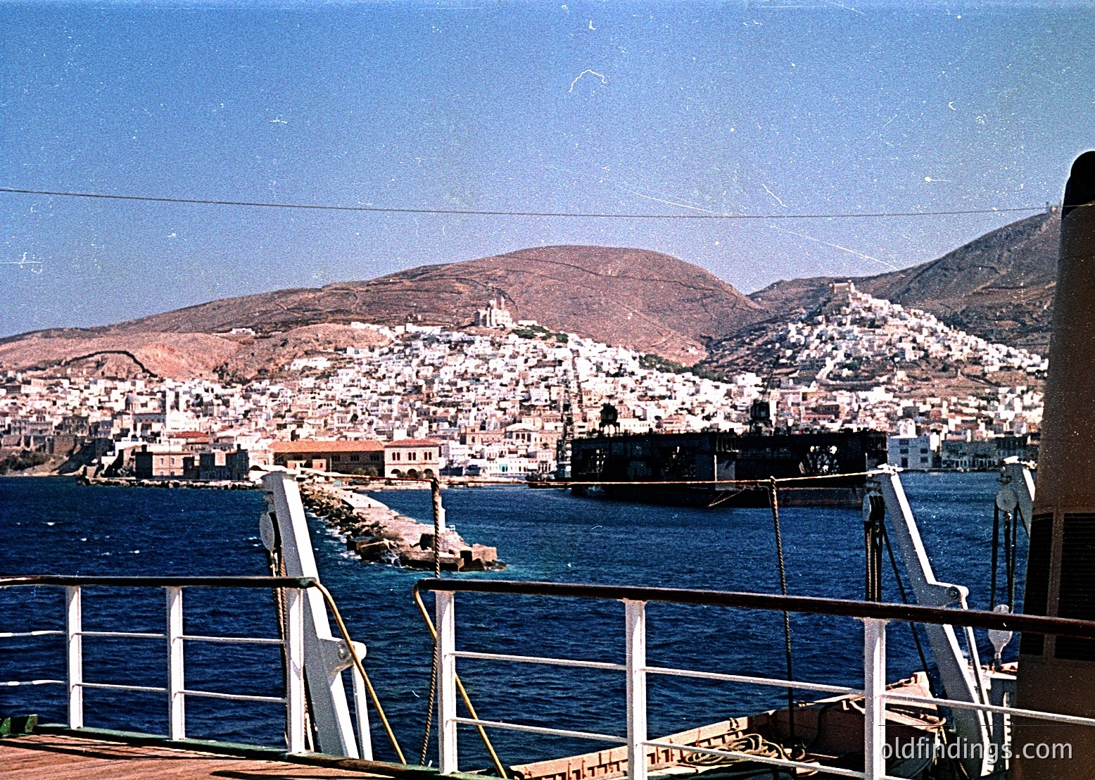 Vintage coastal town nestled on rocky terrain, viewed from a ferry. Dense cluster of whitewashed buildings cascades down hillsides, with a prominent church spire near the center. Deep blue waters frame the foreground, with a visible breakwater extending into the sea. Mid-20th century (1950s–1960s) Mediterranean seascape, likely Greece.