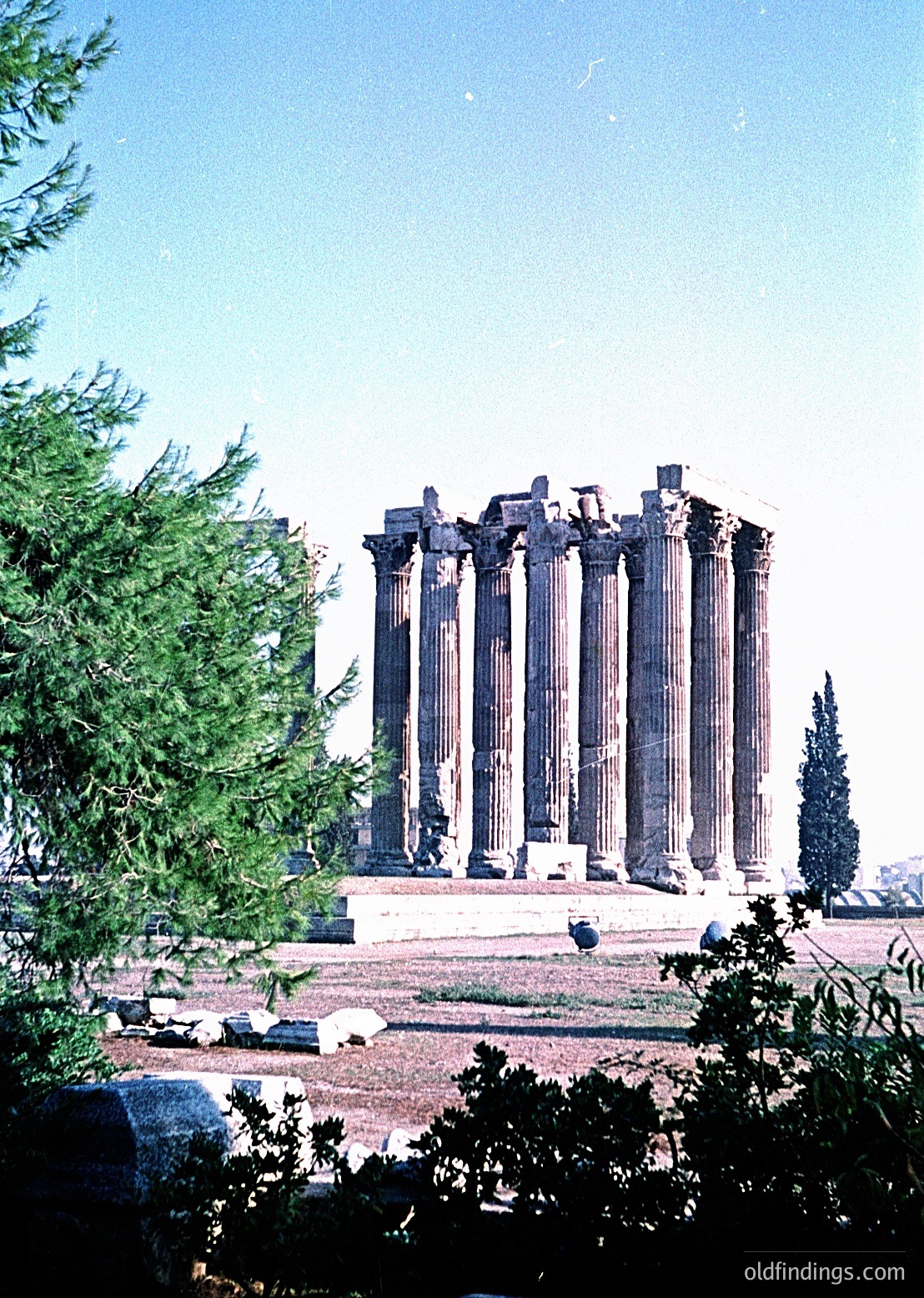 Ancient Greek temple ruins with **16 Corinthian columns**, partially intact, set against a clear sky. Likely the **Temple of Artemis** at Ephesus, Turkey, dating to the **1st century BCE**. Lush greenery frames the structure, emphasizing its grandeur.