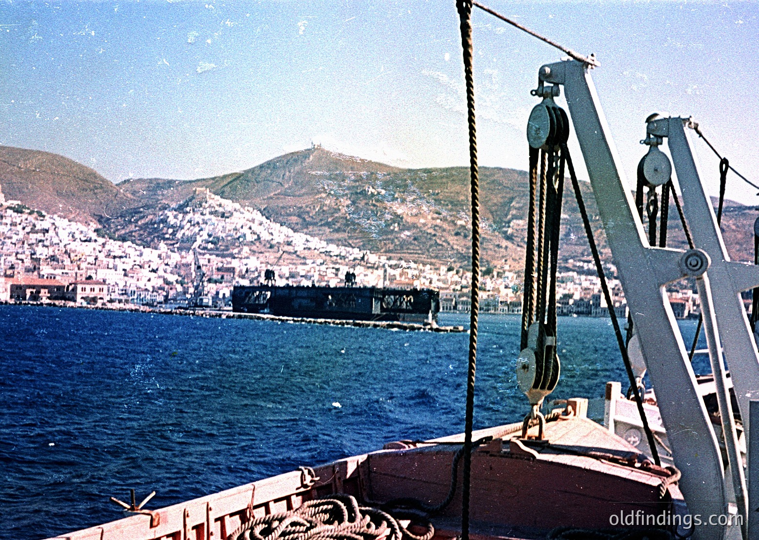 Vintage port scene featuring a mid-20th century cargo ship docked near coastal cityscape. Industrial cranes and ropes dominate foreground, while hills and whitewashed buildings frame the background. Likely Mediterranean due to architecture and lighting.