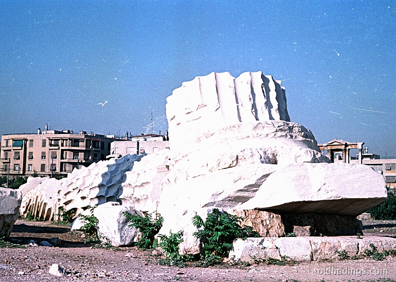 Marble quarry site with stacked, irregularly cut blocks in foreground. Mid-rise residential buildings in background suggest urban-industrial setting. Likely -1980s era due to color and style. Potential research value for and .