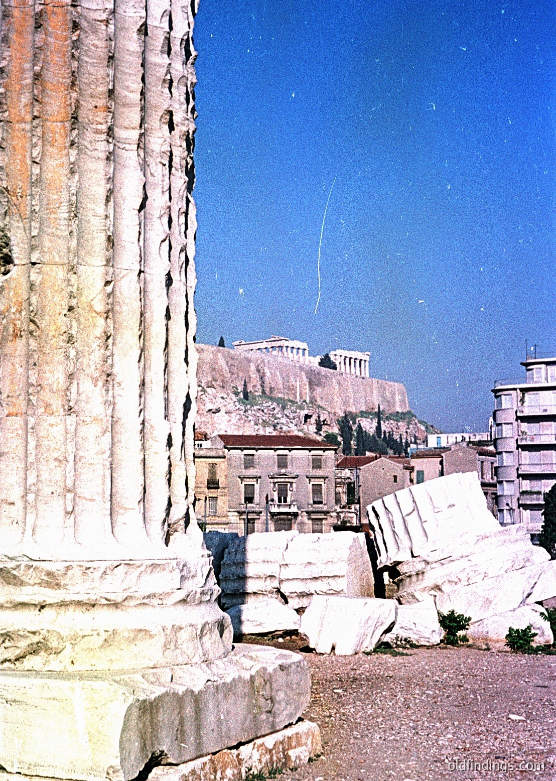 Ancient Greek column fragment in foreground with weathered marble, set against the Acropolis in Athens, Greece. Mid-20th century urban backdrop blends historic ruins with modern buildings. Ideal for historical research, architectural studies, and travel inspiration.