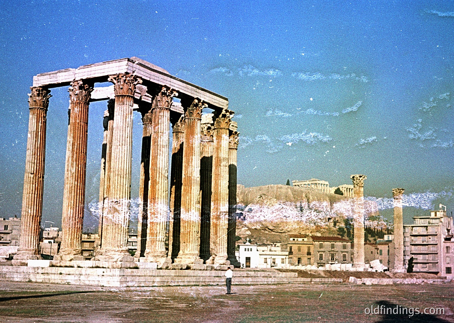 Ancient Doric columns of the Temple of Olympian Zeus in Athens, Greece, with modern buildings in foreground. Mid-20th century sepia-toned photograph captures classical ruins amid urban development.