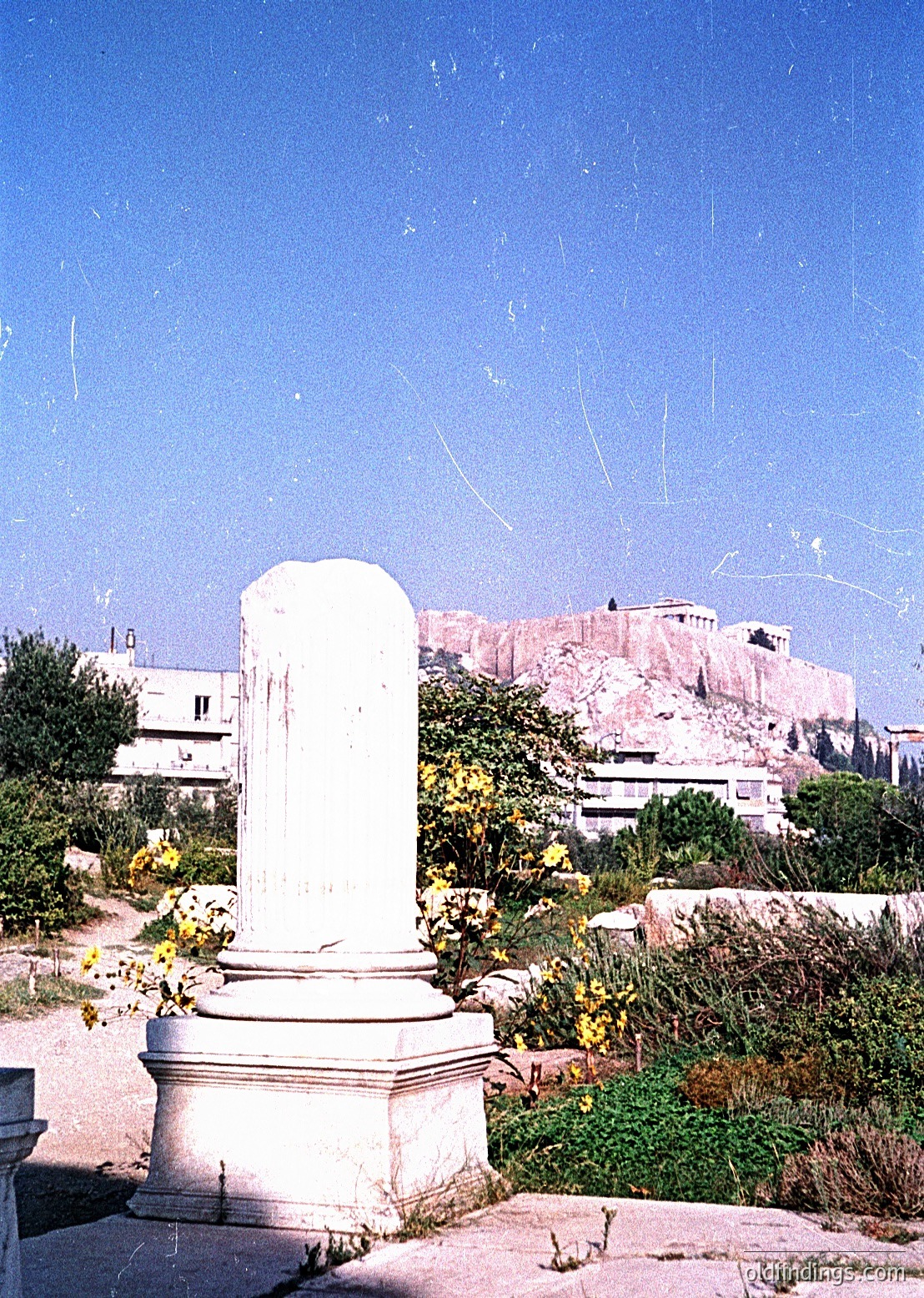 Ancient Greek-style column fragment in a sunlit archaeological park, set against the Acropolis hill in Athens. Weathered marble base with classical fluting, surrounded by sparse vegetation. Mid-20th century preservation efforts evident.