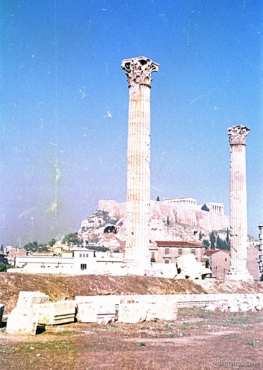 Ancient Greek-style columns with Corinthian capitals standing on a rocky hillside, likely part of the Roman Agora in Athens, Greece. The image captures weathered stone ruins with modern low-rise buildings in the background, suggesting preservation efforts. Circa late 19th to early 20th century based on photographic style.