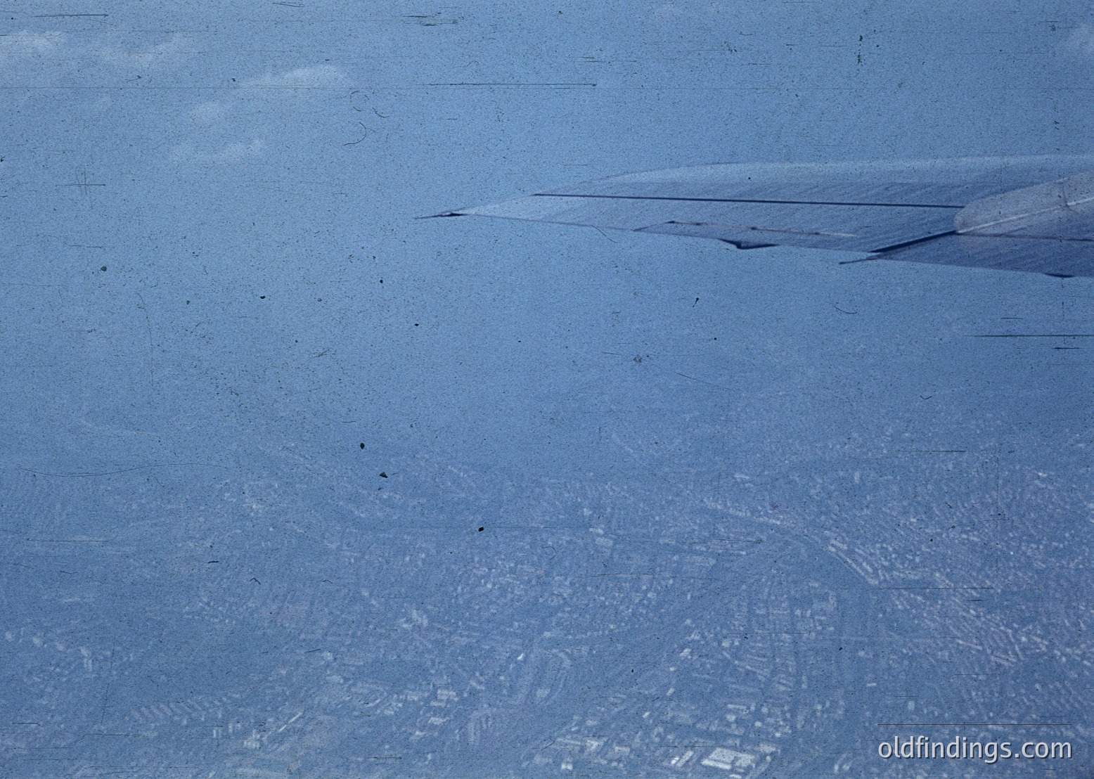 Aerial view of a snow-covered landscape with subtle urban grid patterns beneath. Likely a cityscape obscured by winter snowfall, revealing street layouts and building footprints. The texture suggests compacted snow and urban density.
