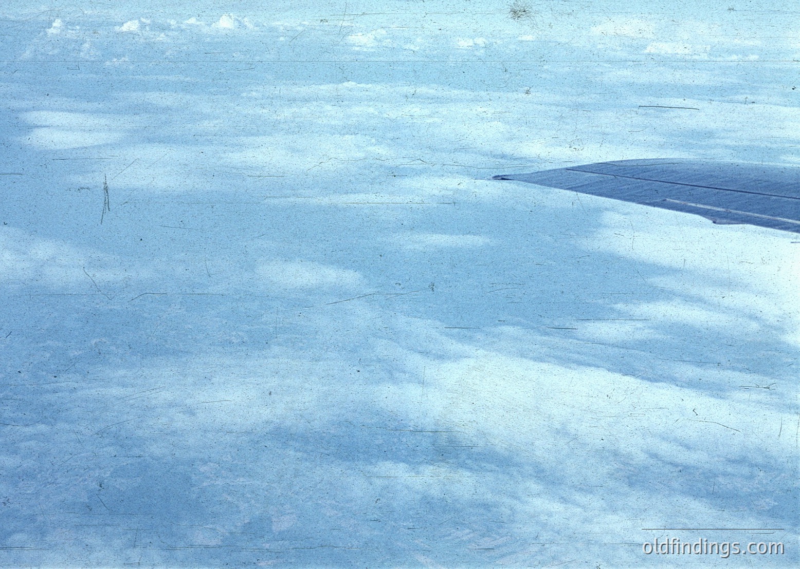 Vintage aerial view of a snow-covered landscape with subtle, wavy terrain patterns. Likely captured during winter, possibly mid-20th century due to film grain and color tone. The distant dark line suggests a road or trail.