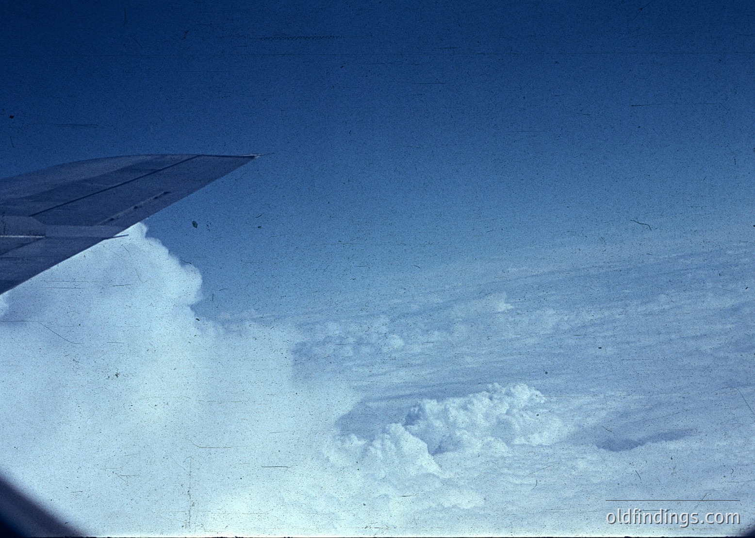 Vintage aerial view of contrails and cloud formations from an aircraft window. The wing edge and condensation trail contrast against the blue sky. Likely captured mid-flight during the 1960s–1980s.