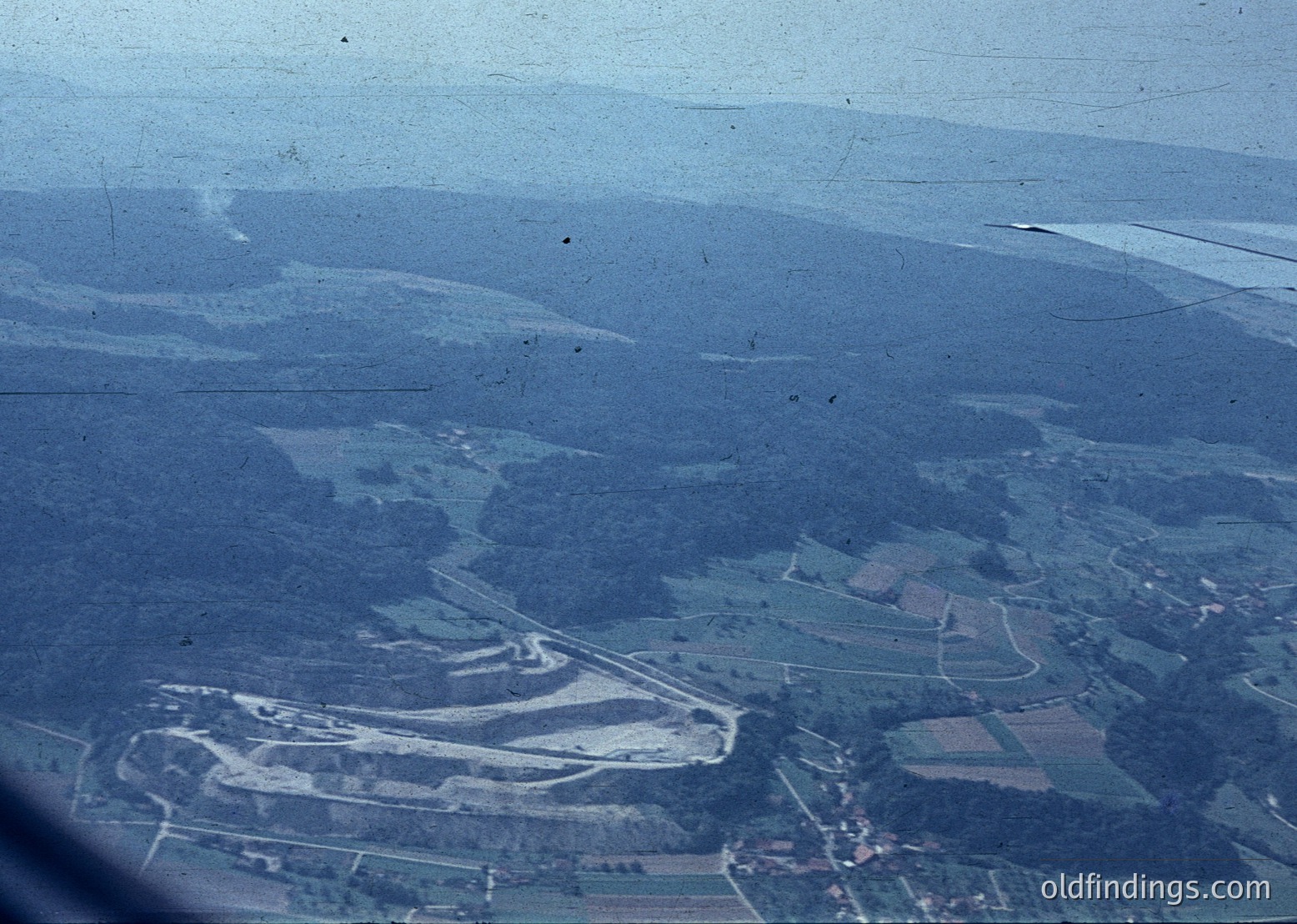 Aerial view of rural terrain with winding roads and agricultural fields, likely mid-20th century. Dense forest patches contrast with cleared farmland, suggesting mixed land use. Vague urban cluster at bottom left hints at nearby settlement.