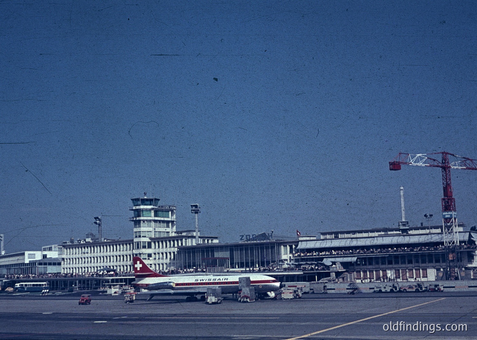 Mid-20th century airport terminal with Swissair jet in foreground. Brutalist architecture with concrete towers and geometric design. Construction crane and vintage vehicles visible. Likely European, 1960s-1970s era.