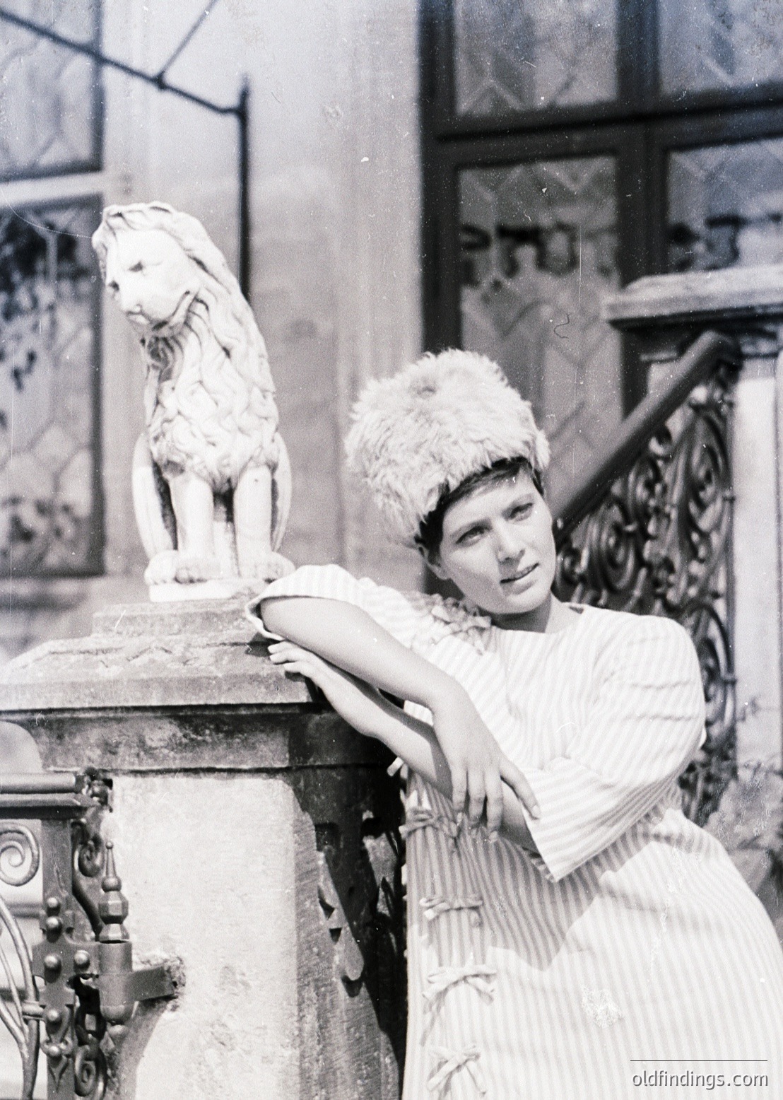 Vintage black-and-white portrait of a woman in early 20th-century fashion, posing beside a marble lion statue atop ornate stonework. Her fur-trimmed hat and draped blouse reflect Art Deco-inspired elegance. Architectural details suggest an urban European setting, likely 1920s–1930s.