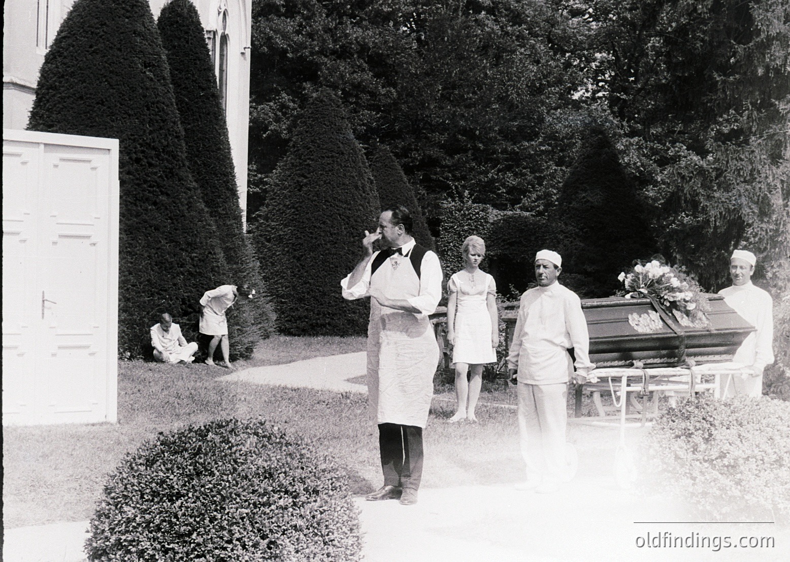 Mid-20th century funeral procession in a manicured garden setting. A man in a chef’s apron and cap stands beside a closed casket draped with floral arrangements, while others in formal attire follow. Whitewashed building and trimmed hedges frame the scene, suggesting a private or institutional location.
