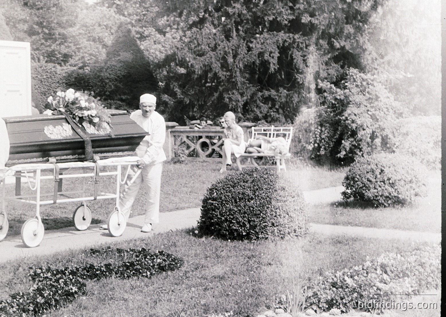 Mid-20th century funeral procession in a landscaped cemetery. A man in formal attire pushes a hearse with floral arrangements, while a child stands nearby. Ornate wrought-iron fence and manicured shrubs frame the scene.
