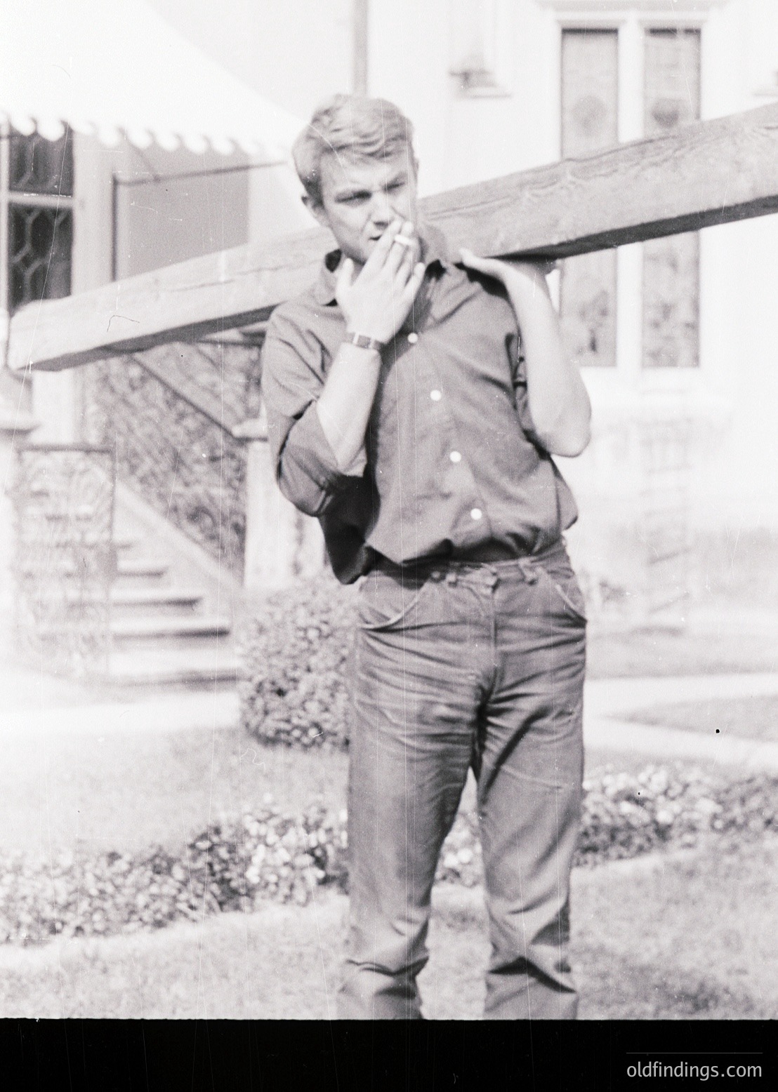 Young man in 1960s-style casual attire—rolled-up sleeves, button-down shirt, and loose-fitting trousers—standing outdoors near a brick staircase and wooden railing. His hand covers his mouth in a candid pose, suggesting a moment of contemplation or laughter. Classic mid-century streetwear and architecture hint at Western European or North American suburban life.