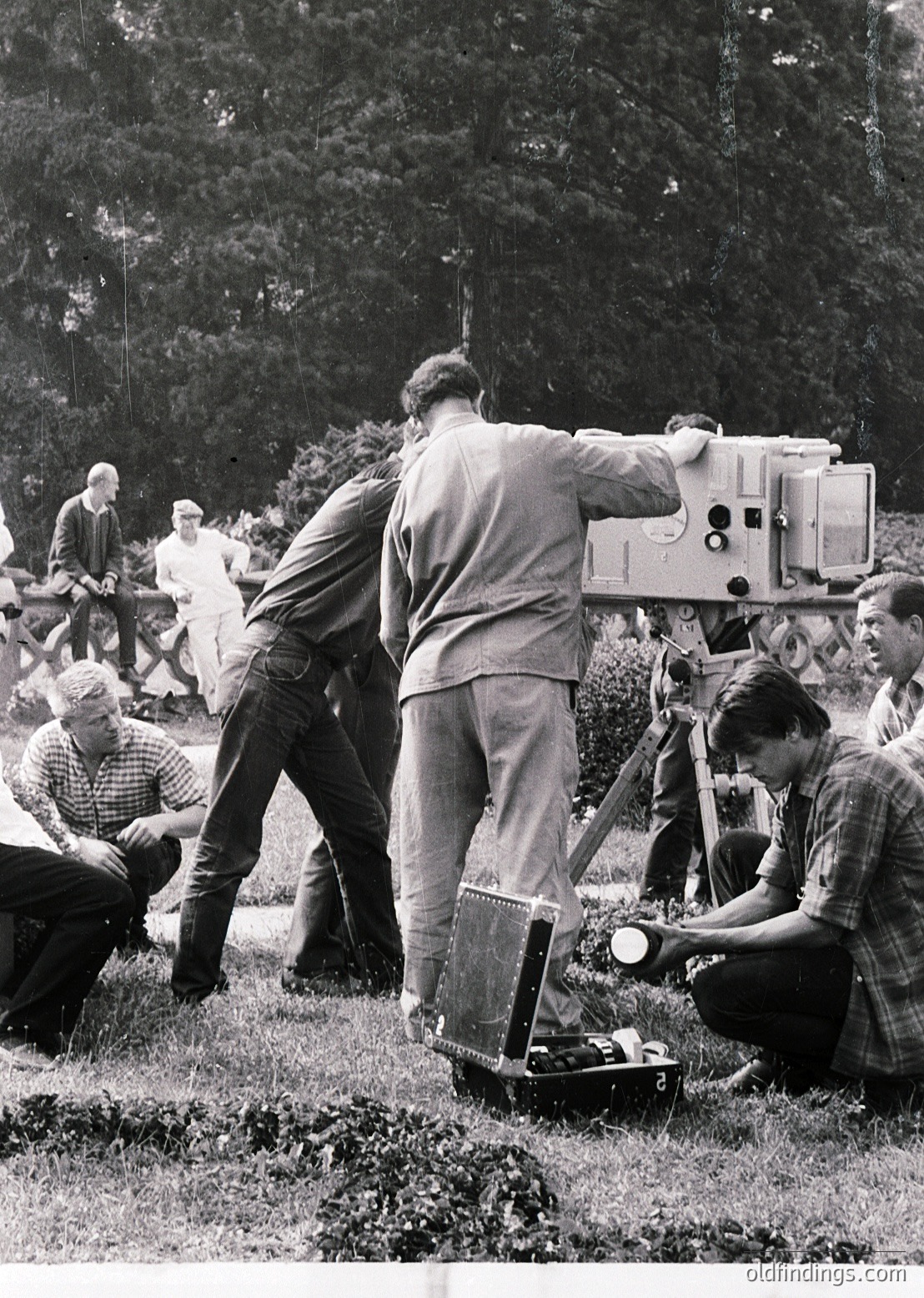 Mid-20th century film crew operating vintage 16mm camera setup outdoors. Crew members in casual 1950s-60s attire—plaid shirts, slacks, and caps—focus on framing shot. Wooden tripod and manual dolly suggest handheld mobility. Lush forest backdrop hints at natural location shooting. Ideal for historical filmmaking research or vintage documentary references.