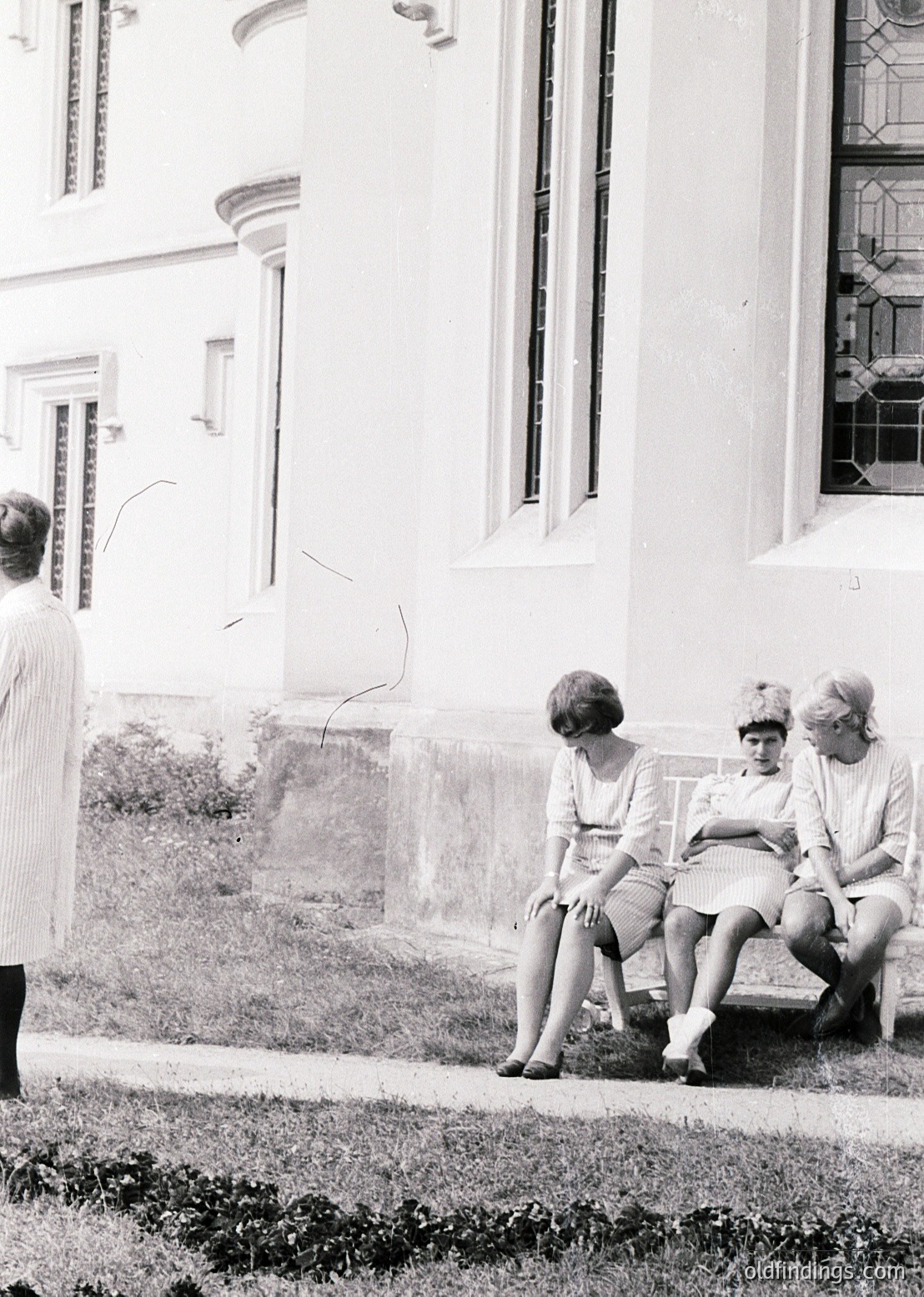 Three women in 1950s-style dresses sit on a low wall beside a grand, classical building with arched windows and decorative moldings. One woman stands nearby, facing away. Mid-century fashion and architecture suggest a European setting, likely for historical or vintage stock use.